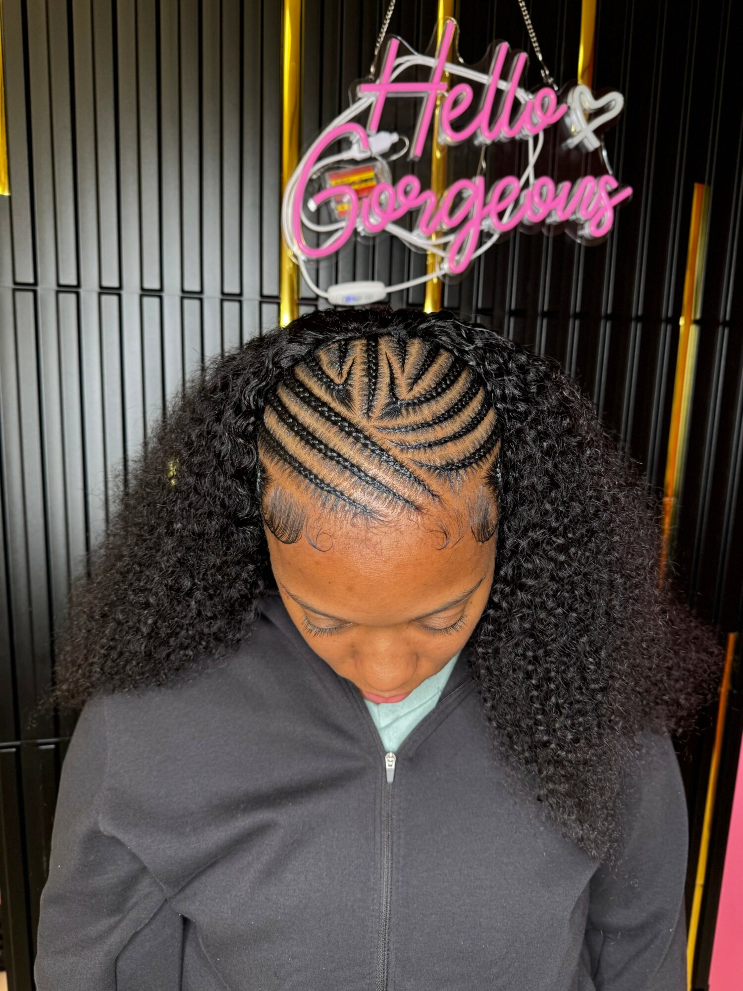 Woman with curly hair and intricate braided hairstyle, standing in front of a black and gold background, with a pink neon sign reading 'Hello Gorgeous' above her head.