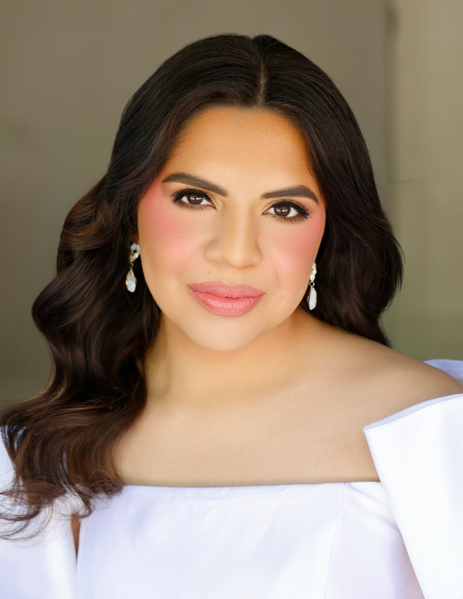 Portrait of a woman with long dark wavy hair, wearing earrings and a white off-shoulder top, smiling softly.