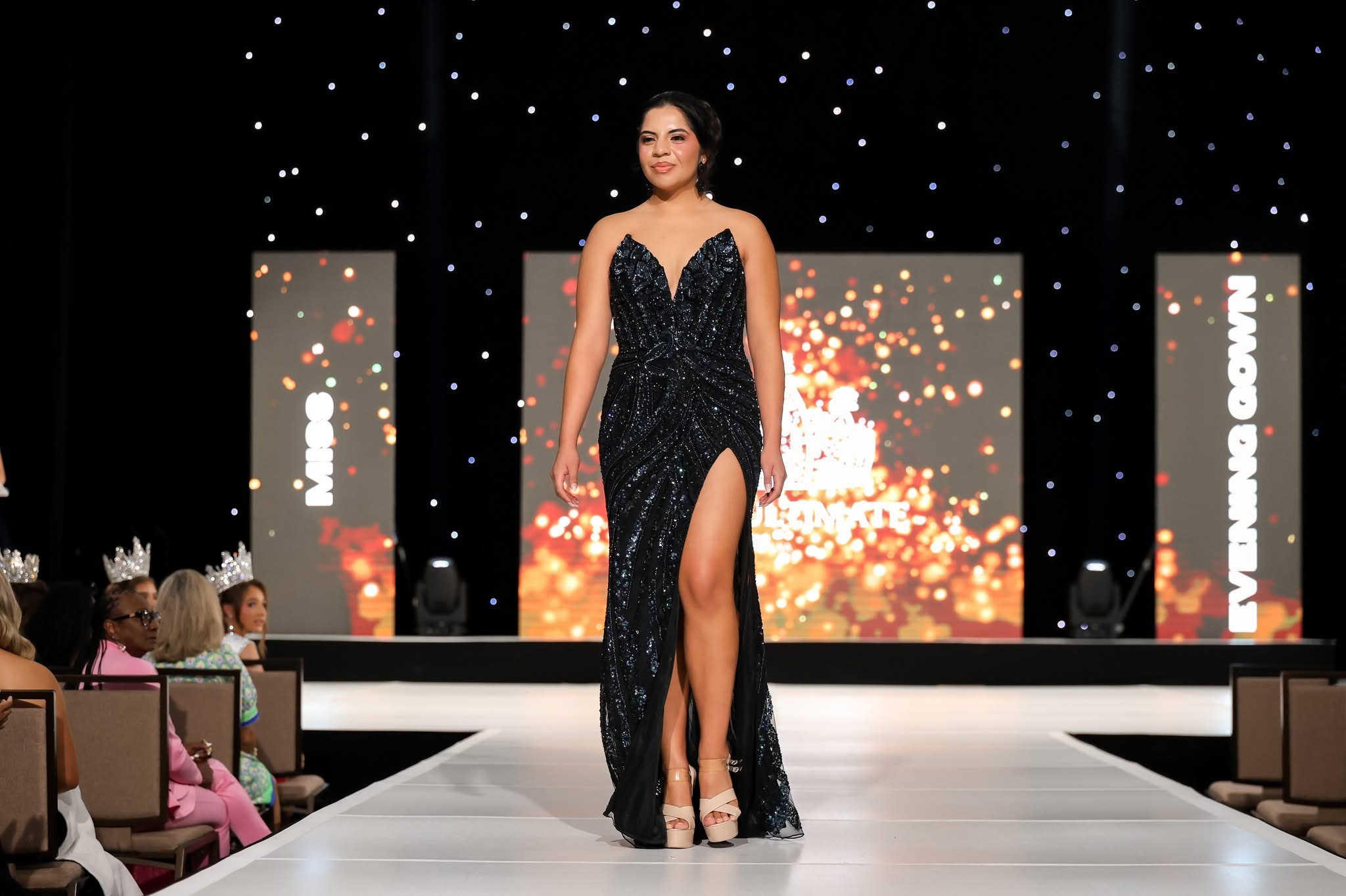 Woman in a black gown with a high slit walking on a runway during a fashion show, audience seated on sides.
