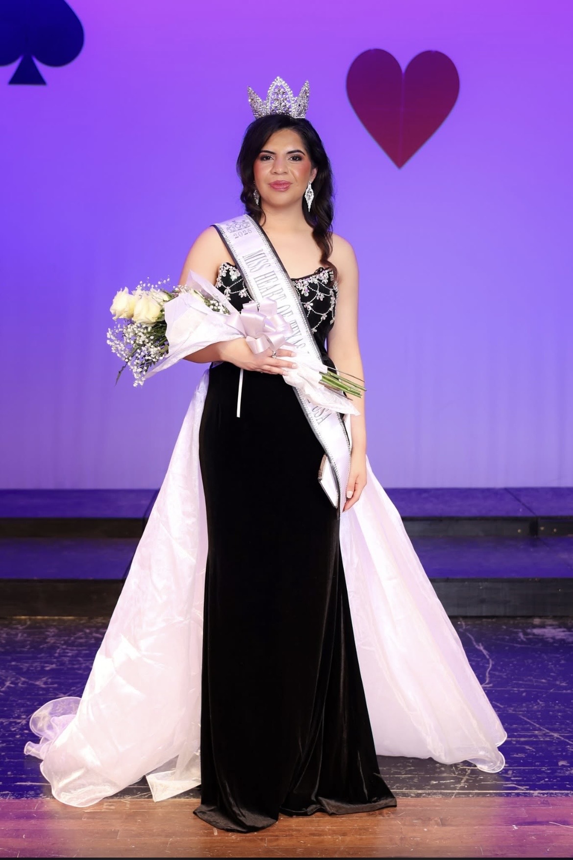 Woman wearing a crown and sash, holding a bouquet, standing on stage with purple background and heart symbols.