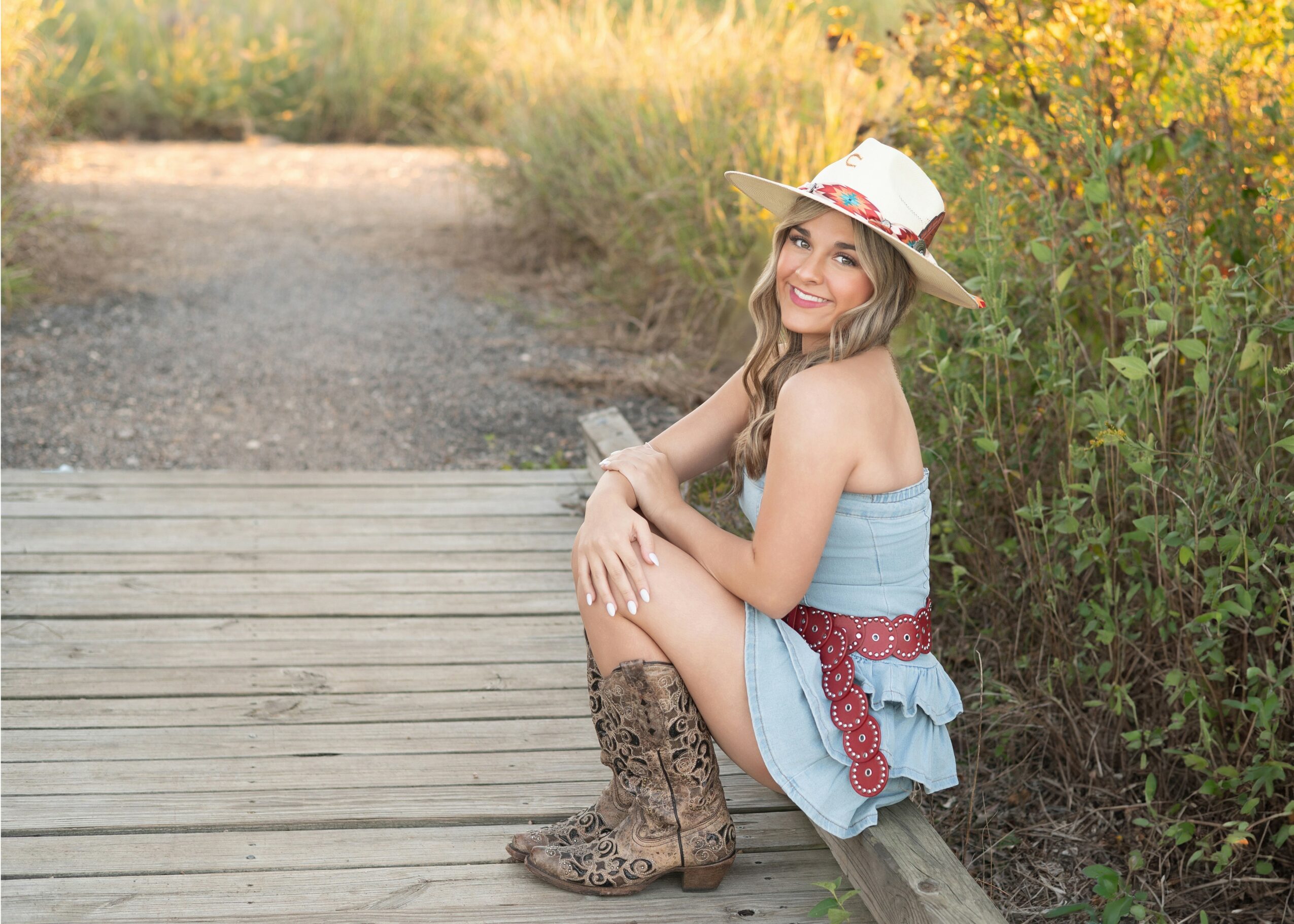 Young woman sitting on a wooden path, wearing a hat, denim dress, and cowboy boots, smiling outdoors with greenery around.