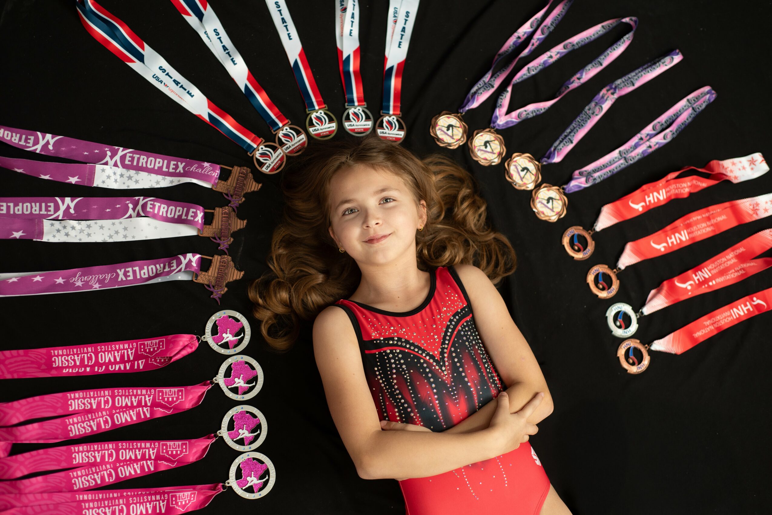 Young girl with curly hair lying on black background, surrounded by medals and ribbons in a circular pattern.