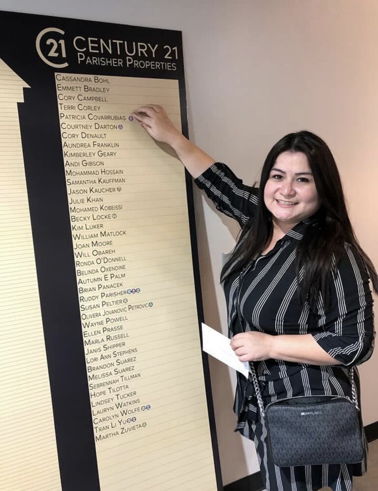 Young woman with long dark hair smiling, pointing at a list on a wall, holding a paper, wearing a striped shirt and carrying a purse.