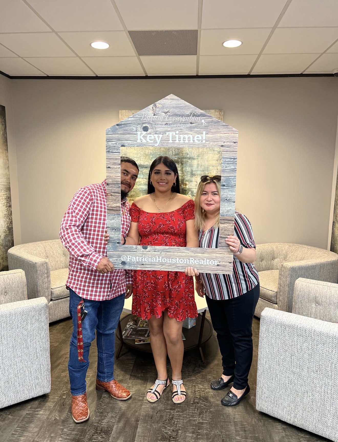 Three people standing together holding a clear rectangular sign, in a room with beige chairs and a decorative backdrop, smiling.