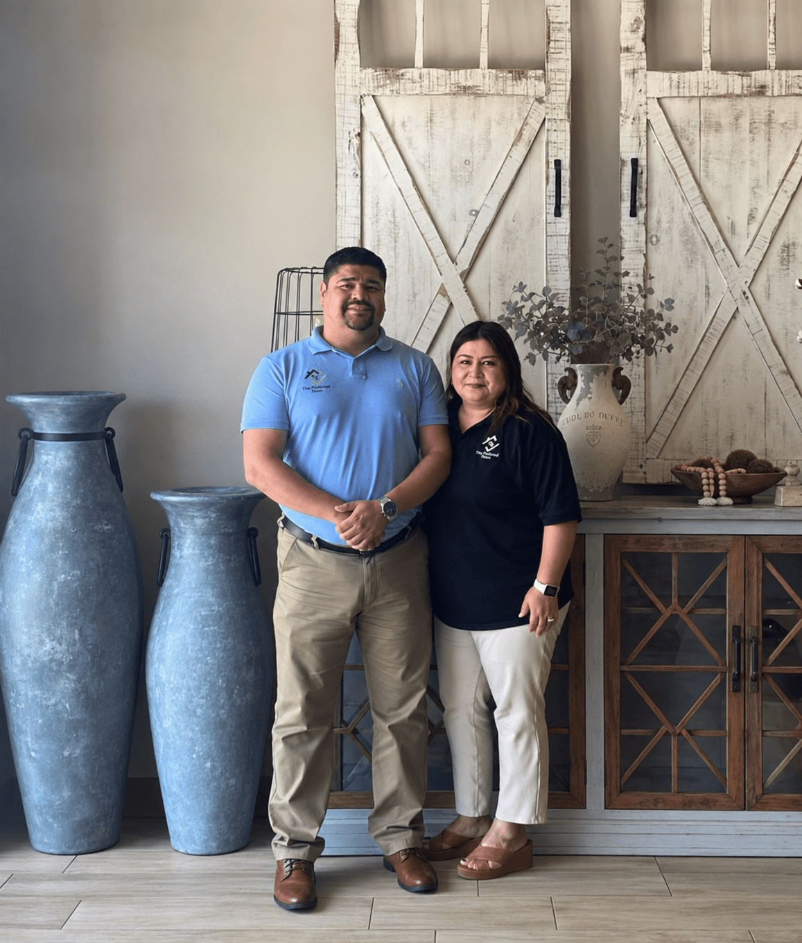 Two people standing indoors, smiling, with large vases and a wooden cabinet behind them.