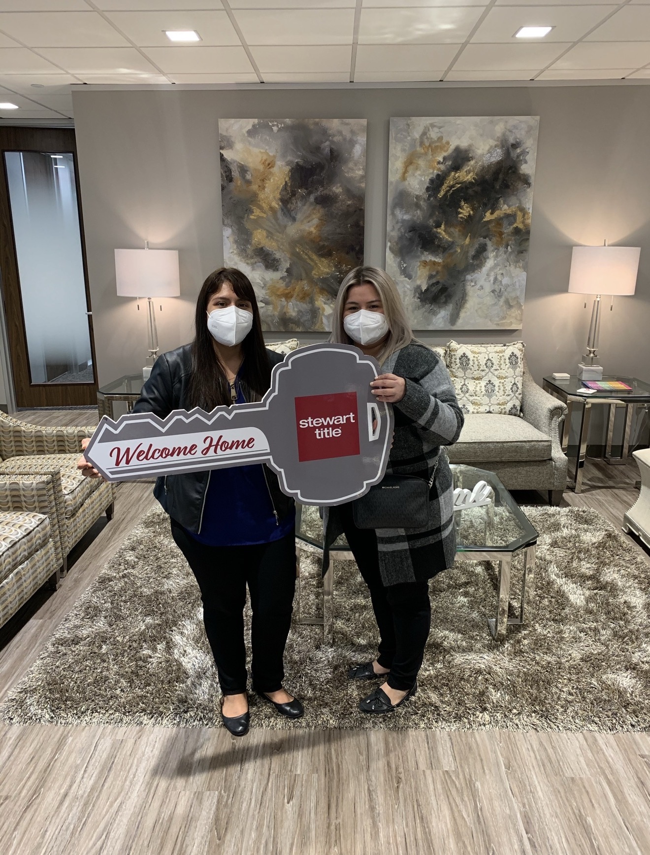 Two women wearing masks stand in a living room holding a sign and a mug-shaped cutout, with artwork and lamps behind them.
