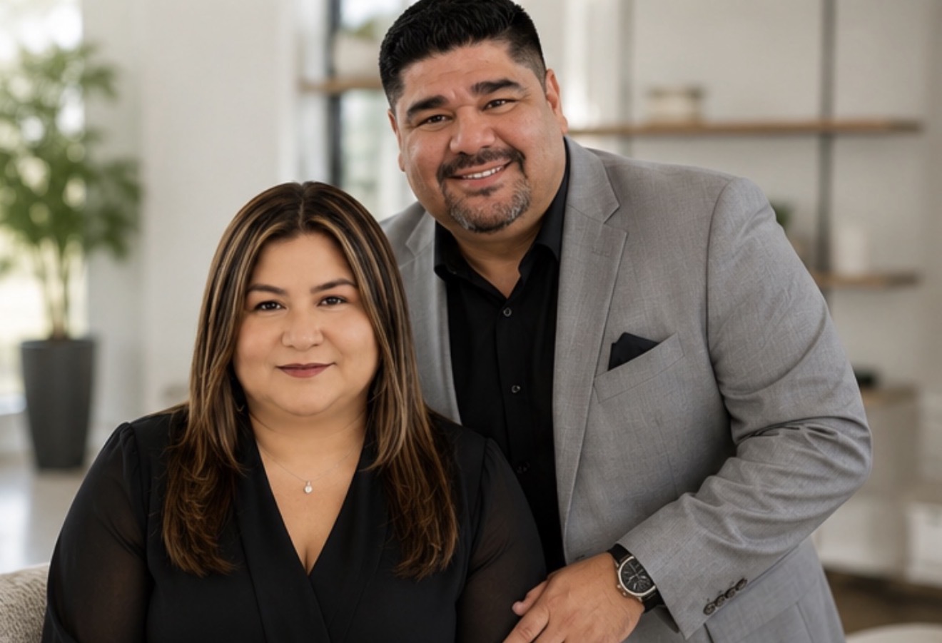 Two people, a woman and a man, smiling indoors with shelves and a plant in the background.