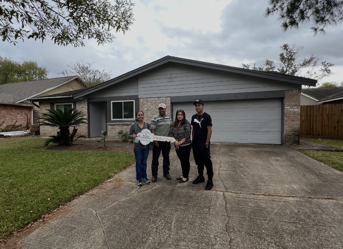 Four people stand in front of a house with a two-car garage, holding a large key, on a driveway with a lawn.