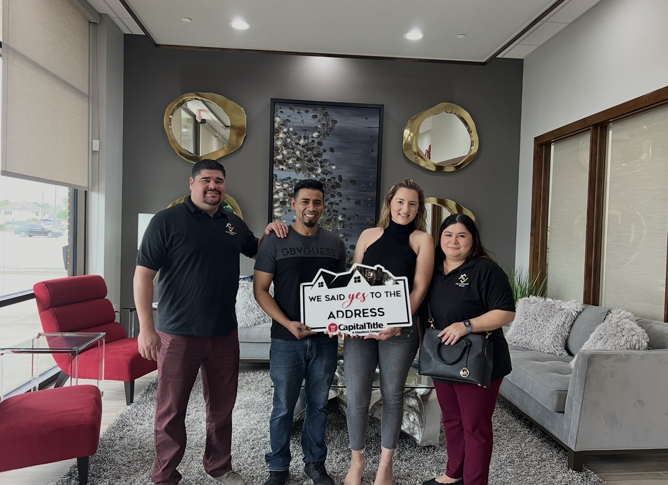 Four people standing in a room holding a sign, with a gray wall, two round mirrors, and a gray sofa behind them.