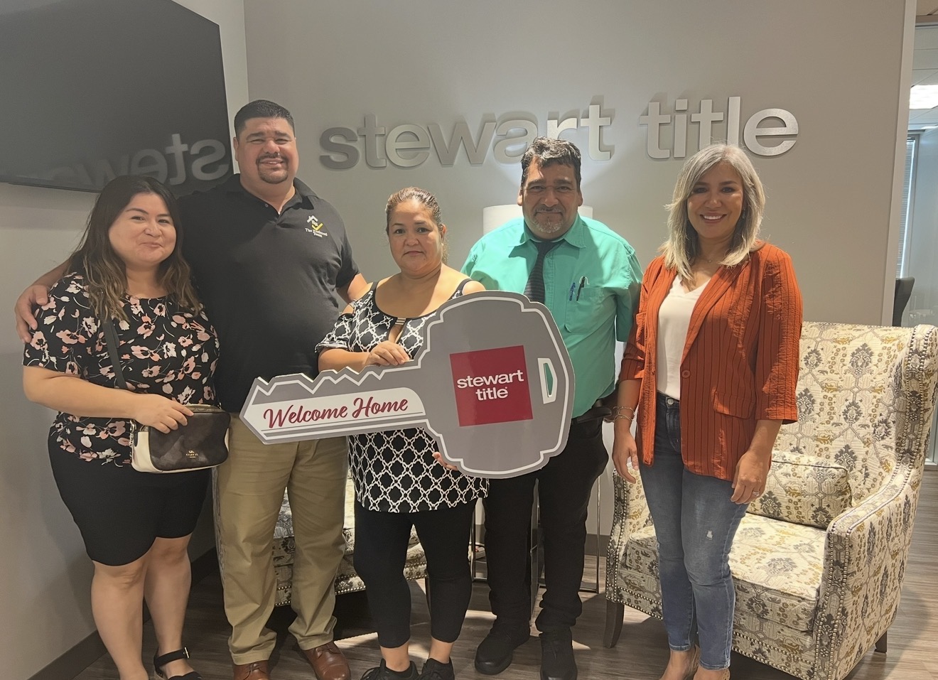 Group of five people standing indoors, holding a welcome sign, smiling, with a patterned armchair nearby.