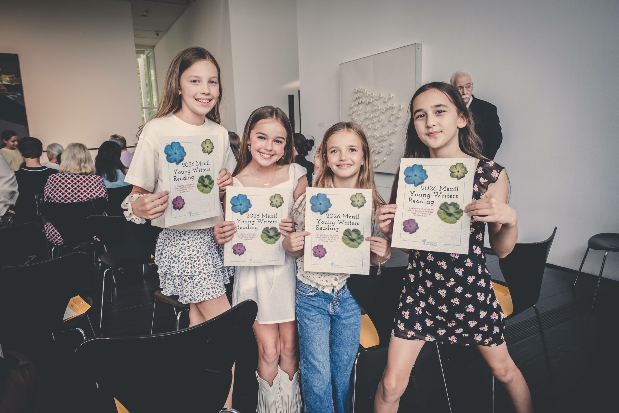 Four young girls holding certificates, smiling, in a room with seated people and a white wall background.