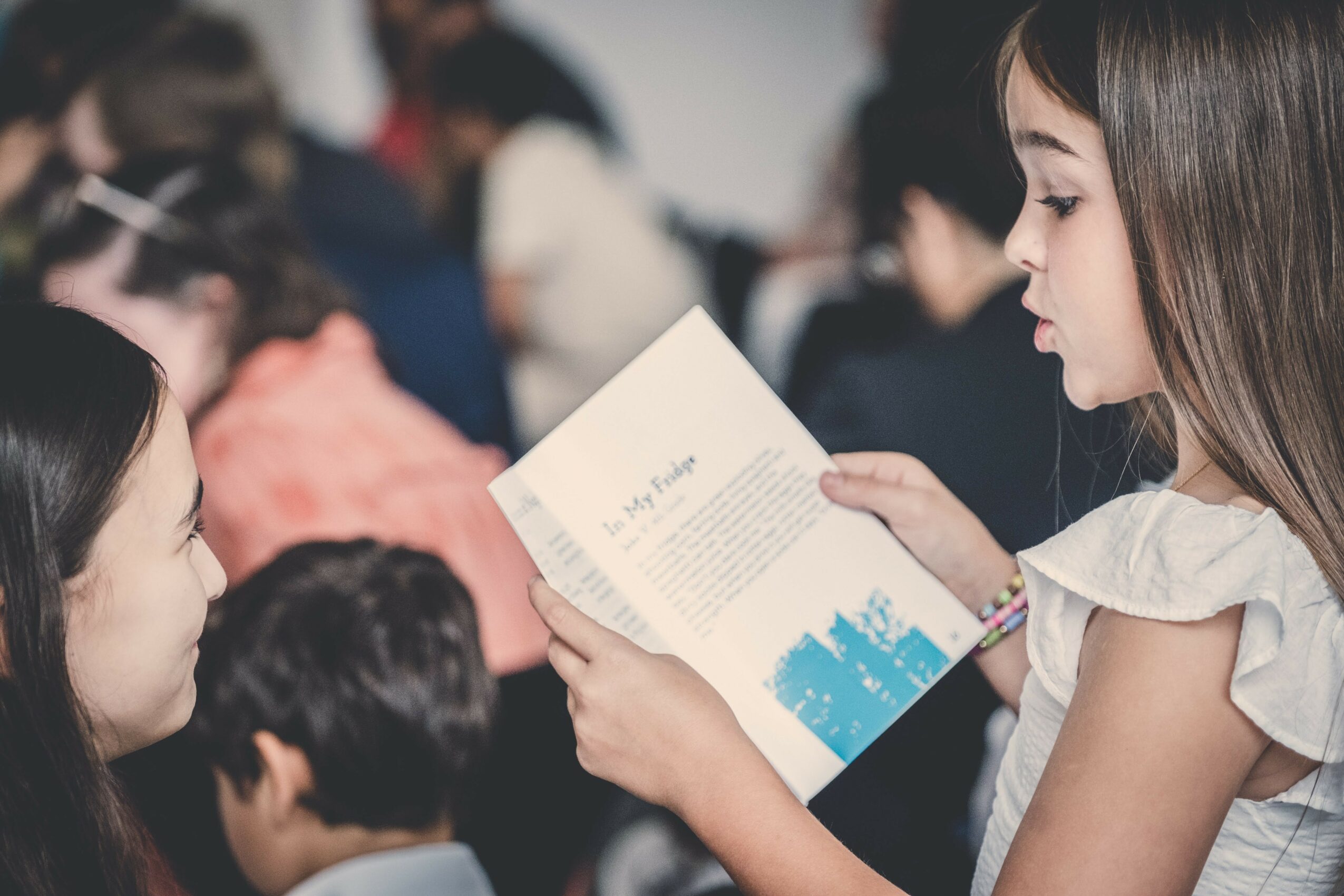 Girl reading a booklet in a crowded indoor setting, with other people in the background.