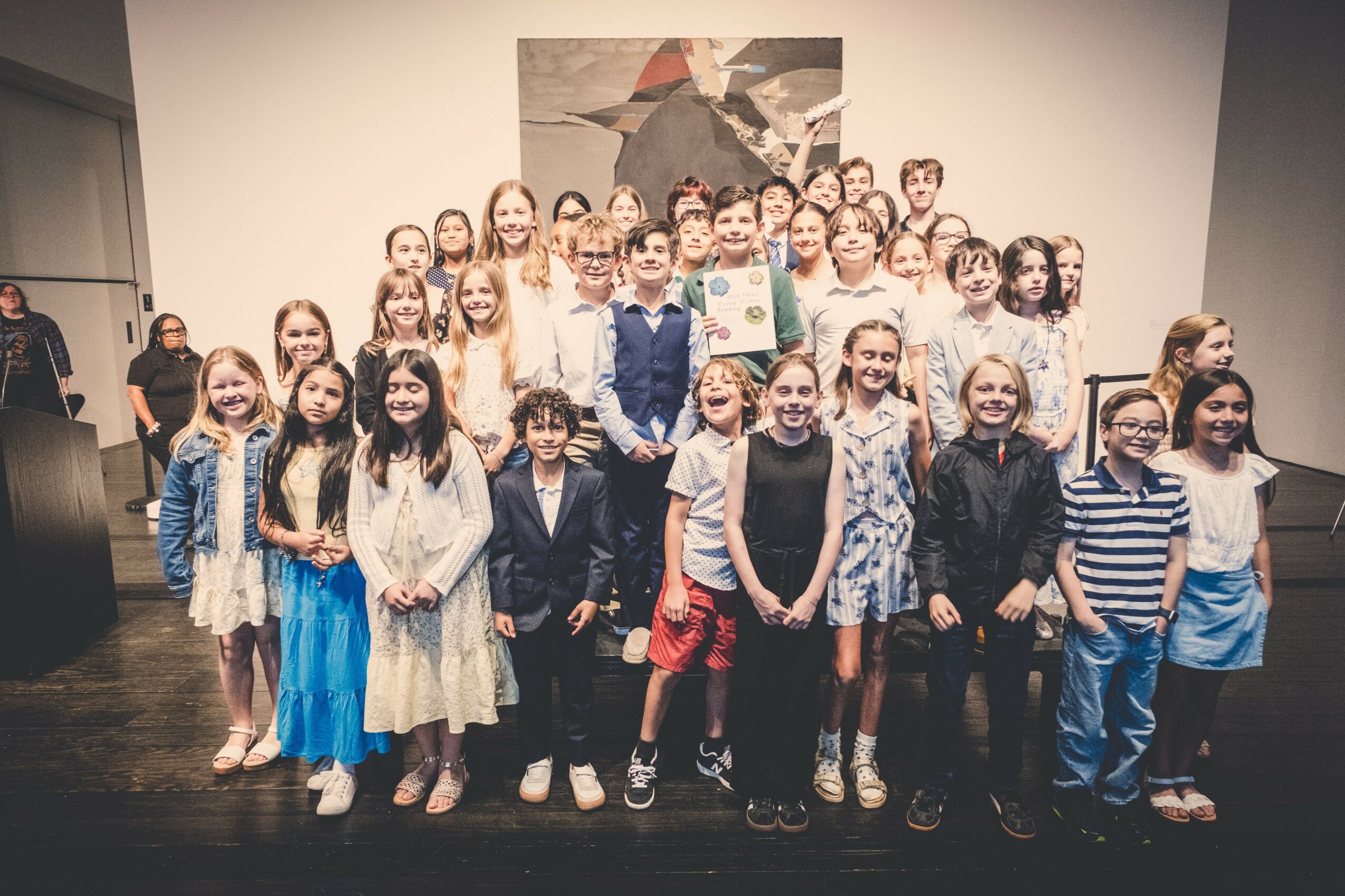 Group of children and adults standing together in a room with a large abstract painting on the wall behind them.