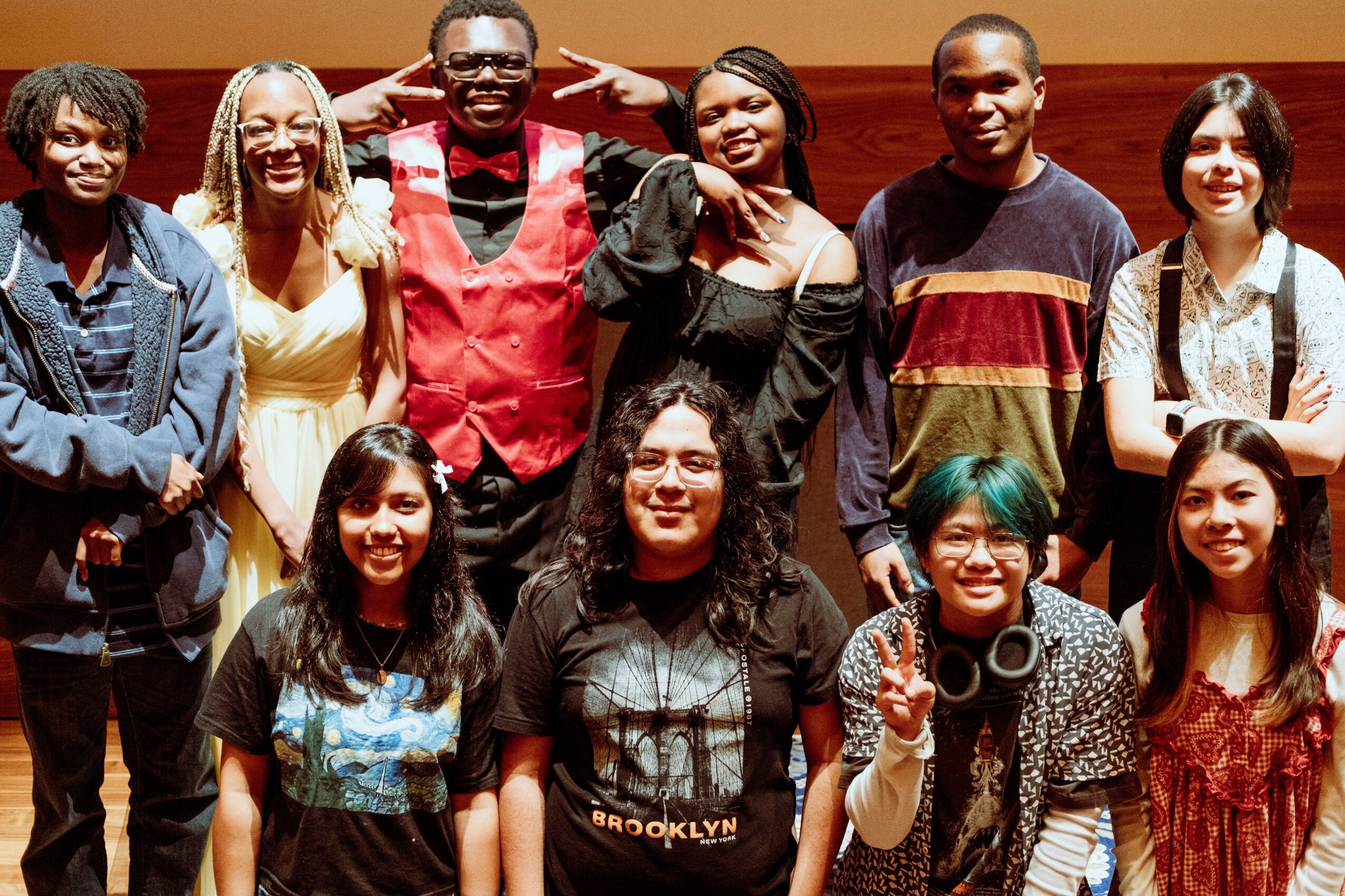 Group of ten diverse young people smiling, standing together indoors, some making peace signs, in casual and stylish clothing.