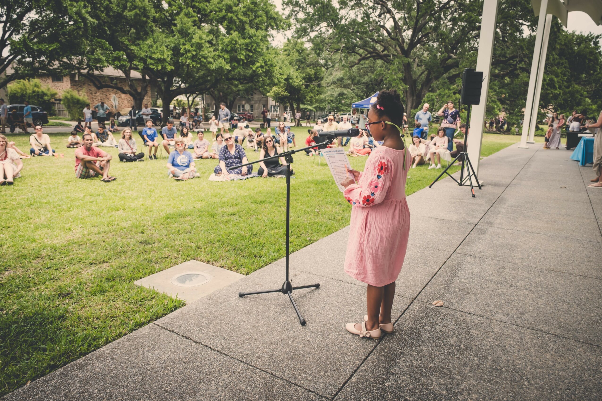 Girl in pink dress speaking into microphone outdoors, audience seated on grass, trees in background.