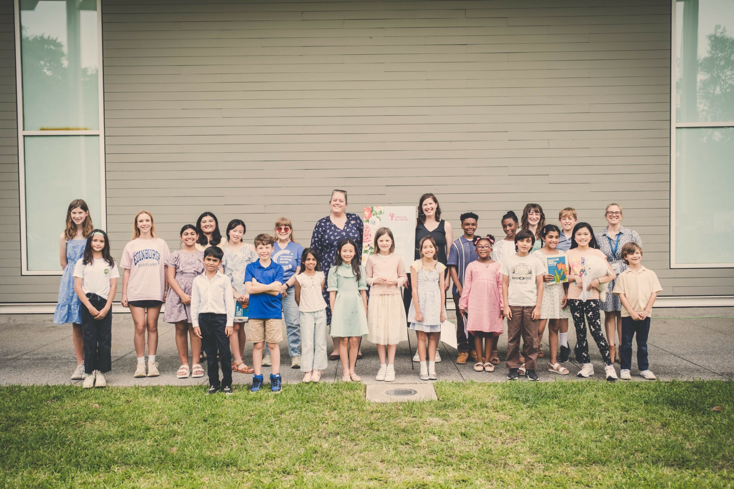 Group of children and adults standing outside in front of a large closed garage door, with grass in the foreground.