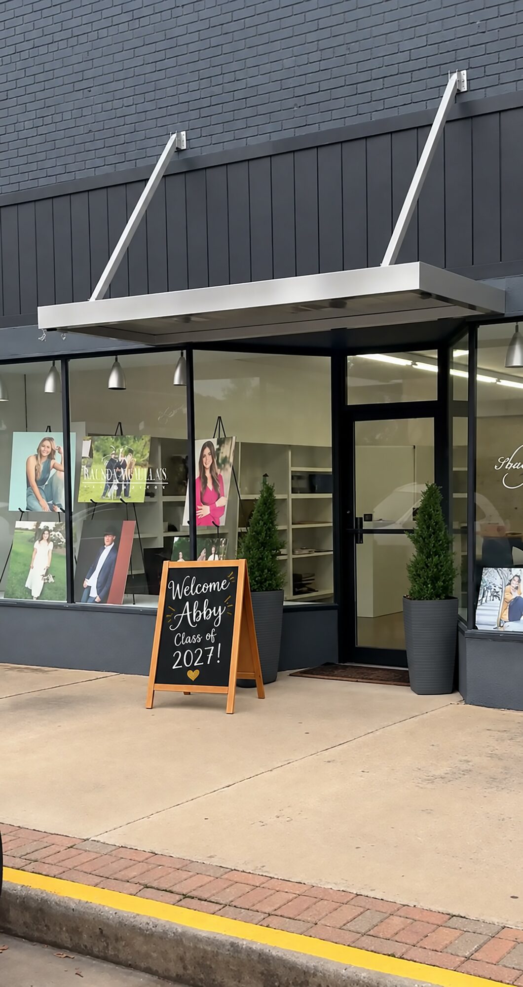 Storefront with large glass windows, a black door, and a chalkboard sign outside, with potted plants on either side.