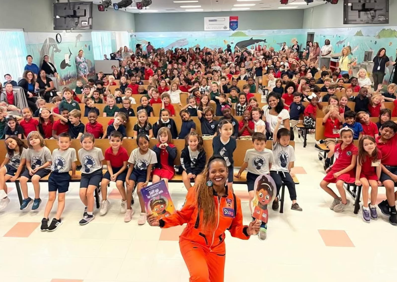 Large group of children seated in a hall with a woman in orange standing in front, holding books and smiling.