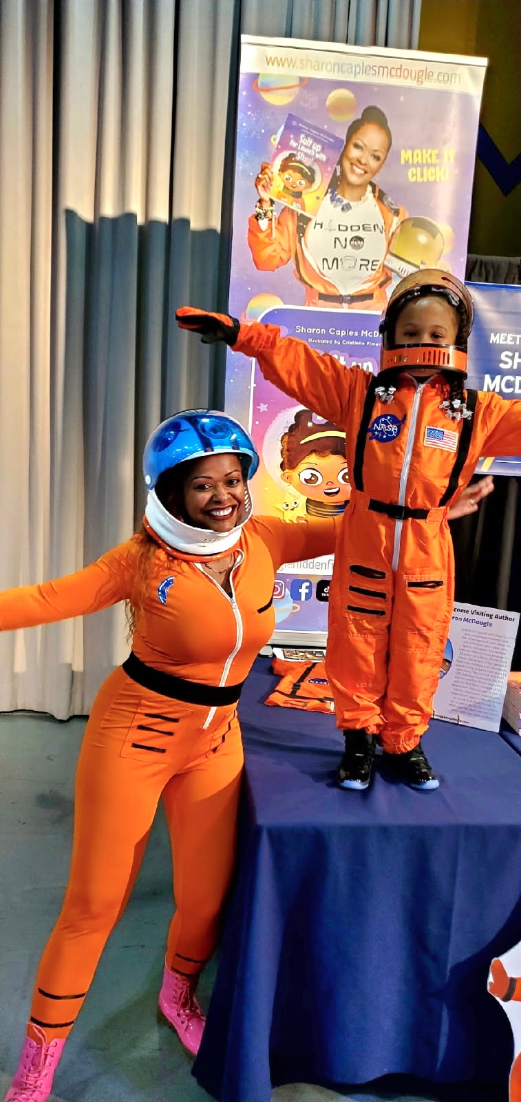 Two children in orange astronaut costumes, one standing on a table and the other pointing, with a colorful poster behind them.