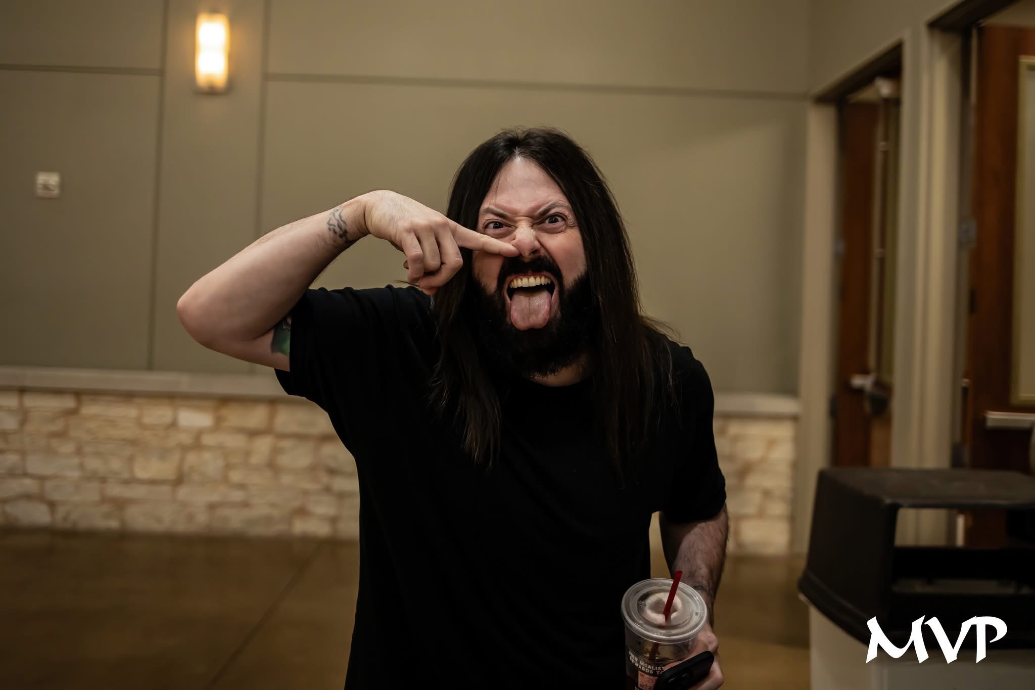 Person with long dark hair and beard making a fierce face, pointing at their eye, holding a drink, indoors.
