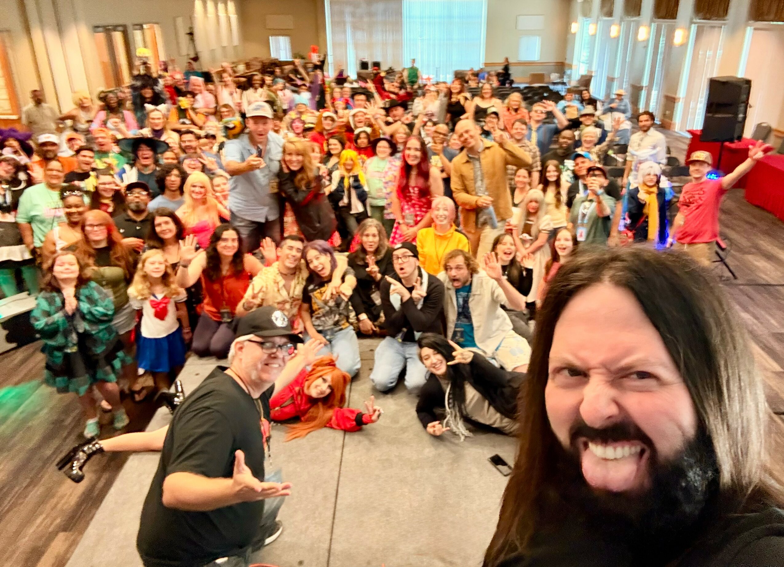 Large group of people gathered indoors, smiling and posing for a selfie, with a person in the foreground taking the photo.