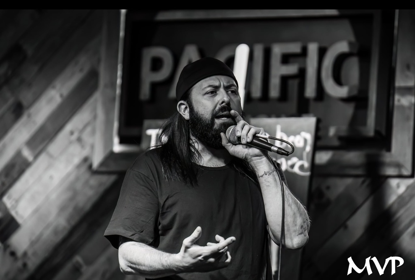 Man with long hair and beard singing into a microphone on stage, background shows a sign with partially visible text.