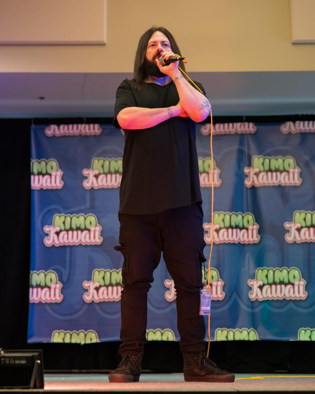 Person with long hair and beard holding microphone on stage with colorful backdrop, wearing black shirt and pants.