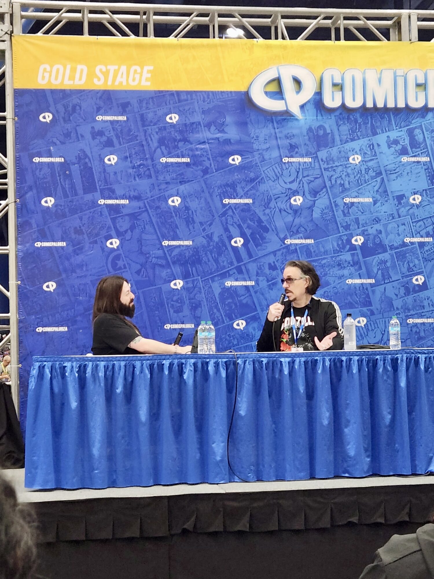 Two people sit at a table on stage with a blue backdrop and a yellow banner reading 'GOLD STAGE'.