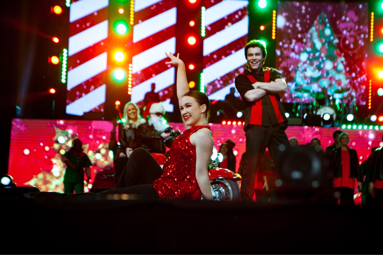 People on stage with Christmas decorations and colorful lights, some holding gifts, festive atmosphere.
