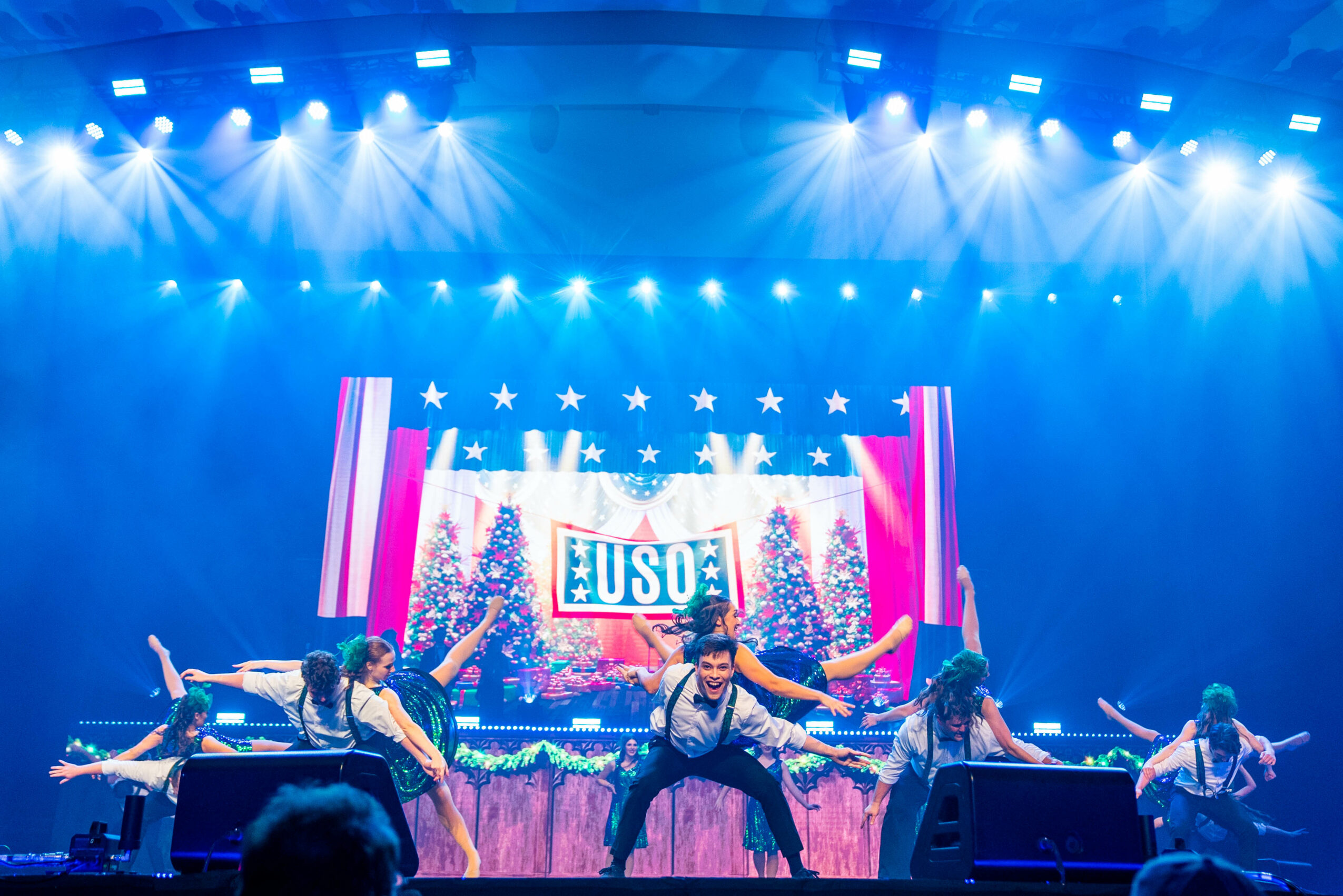 Group of performers dancing on stage with a patriotic backdrop and bright stage lights.