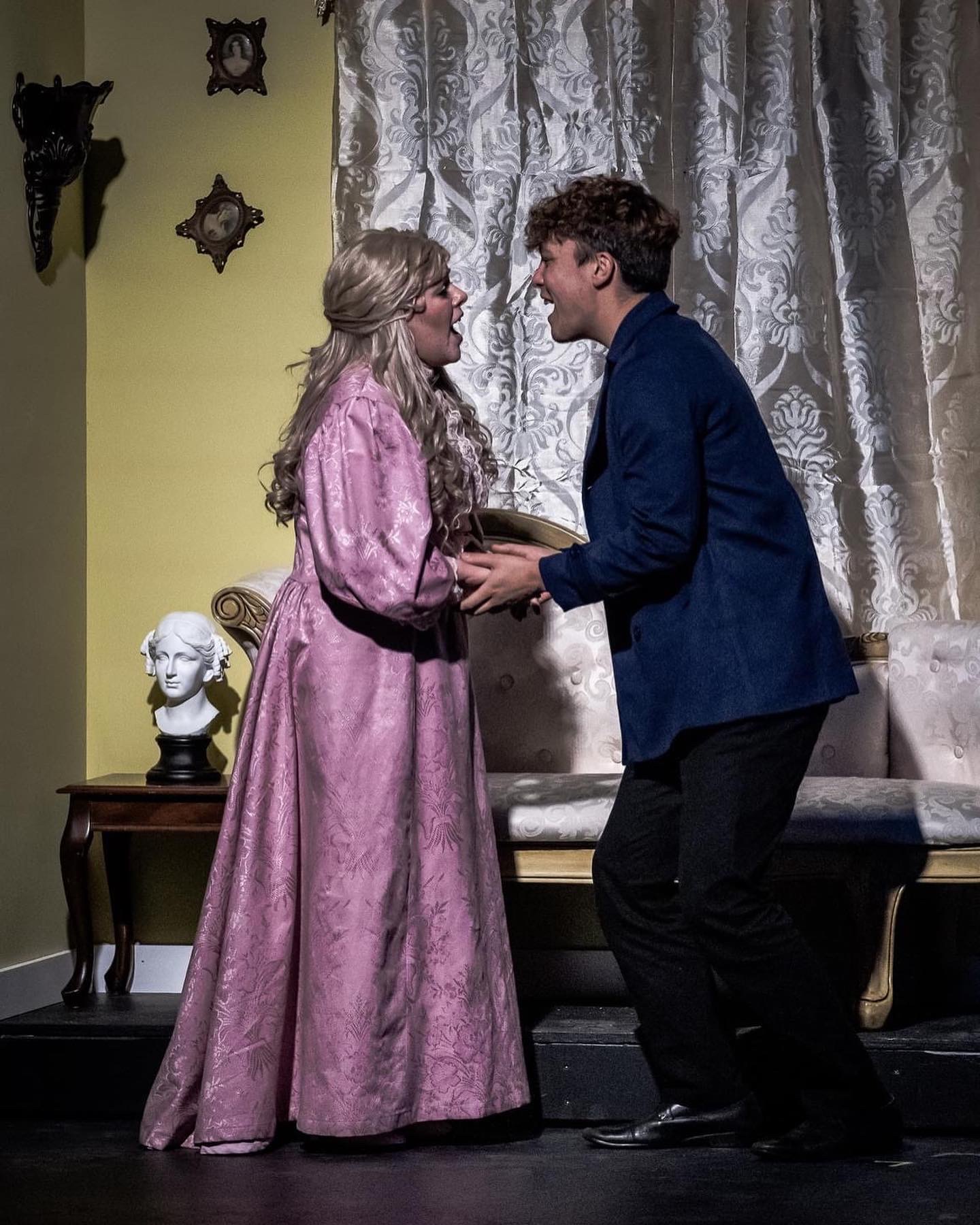 Two people, a woman in a pink dress and a man in a blue jacket, face each other indoors with a curtain behind them.