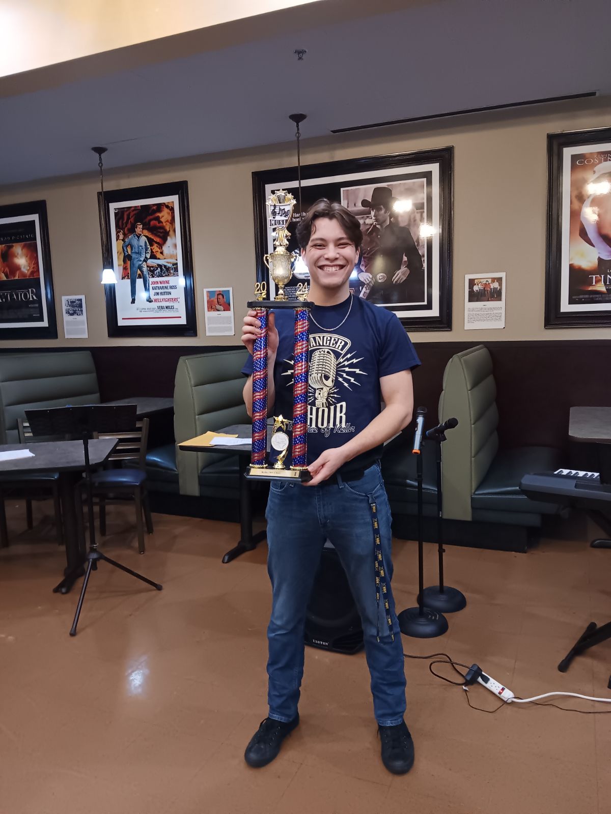 Young person holding a tall trophy, smiling, standing in a room with framed pictures on the wall.