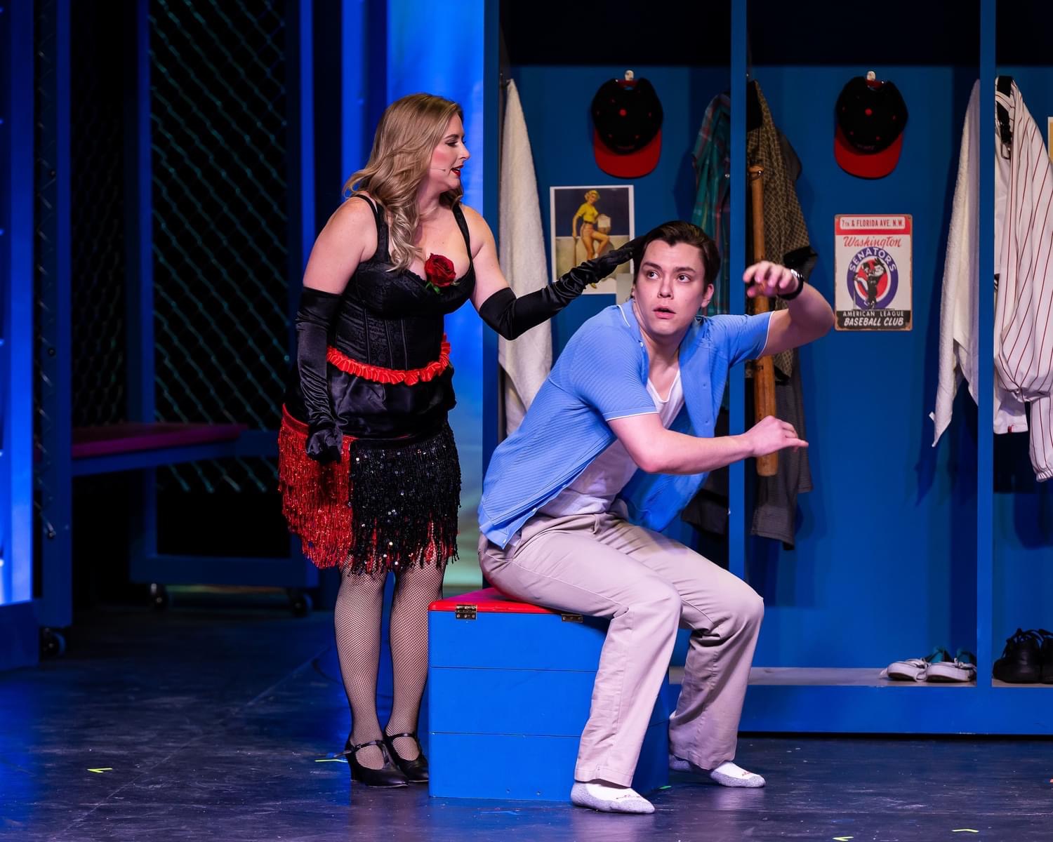 Two people in a locker room, one sitting on a bench and the other standing, with lockers and clothing in the background.