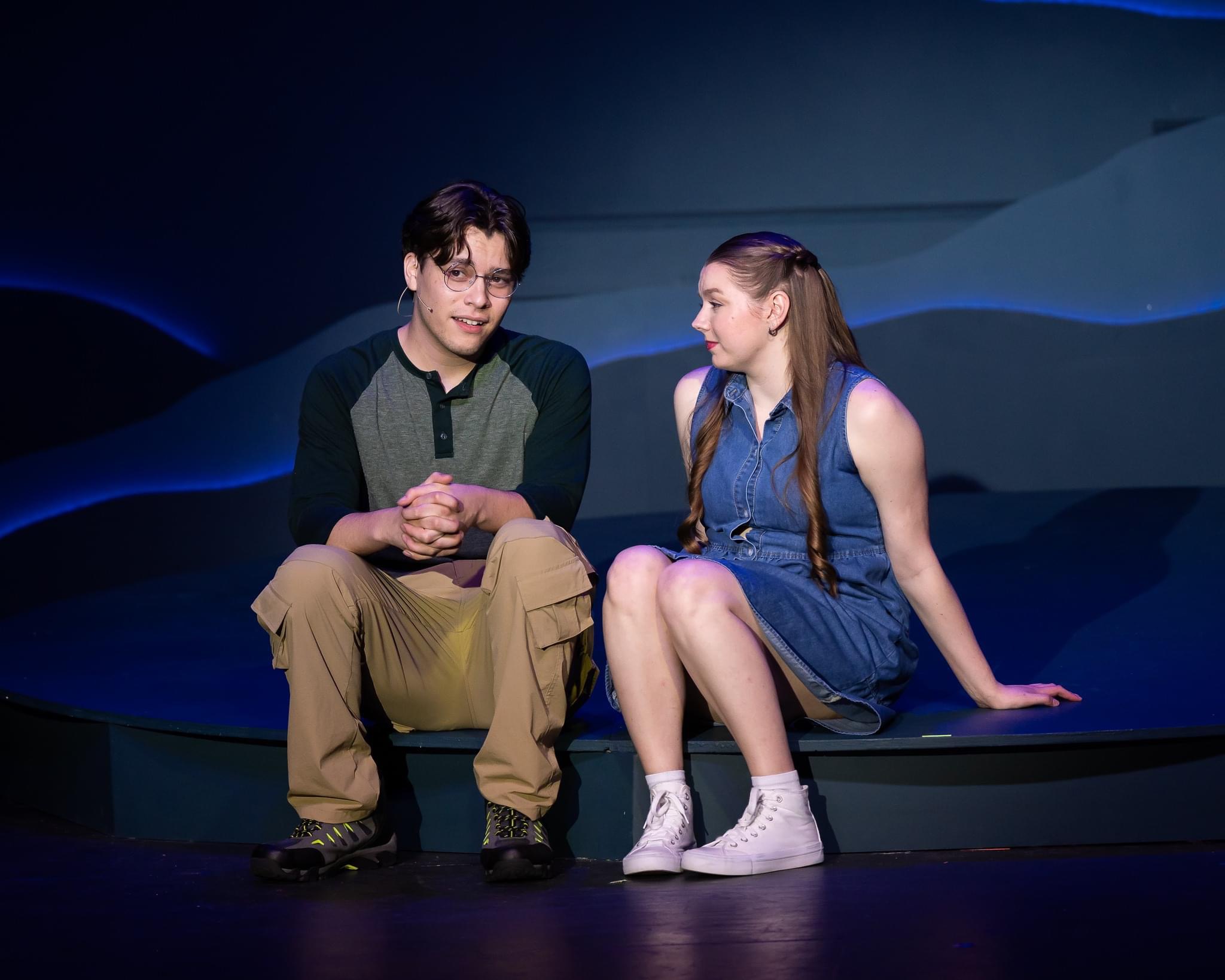 Two young people sitting on a stage, one male and one female, engaged in conversation against a dark background.