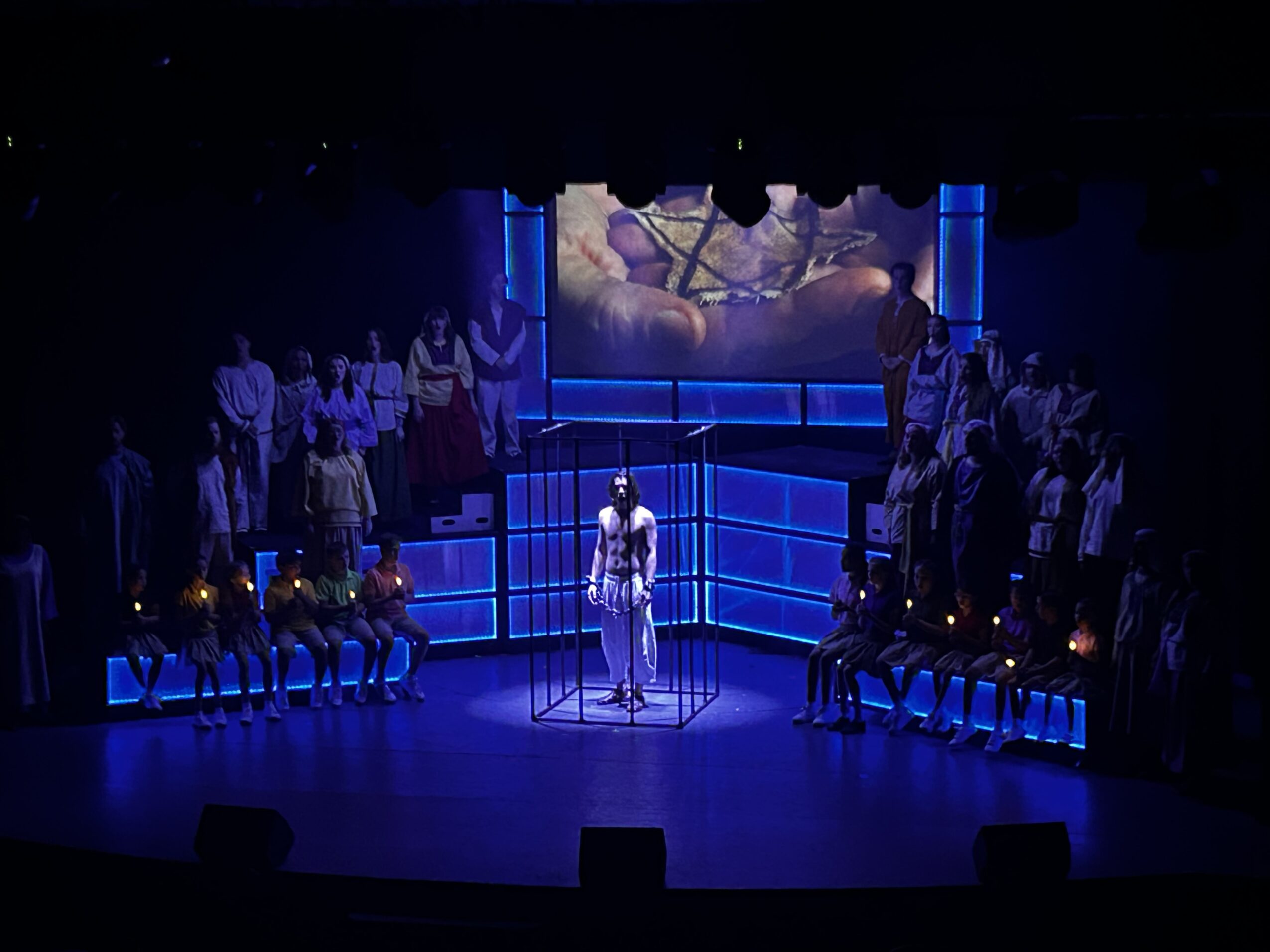 Stage with a person standing at a microphone, surrounded by seated and standing people, illuminated by blue lighting, with a large screen above.