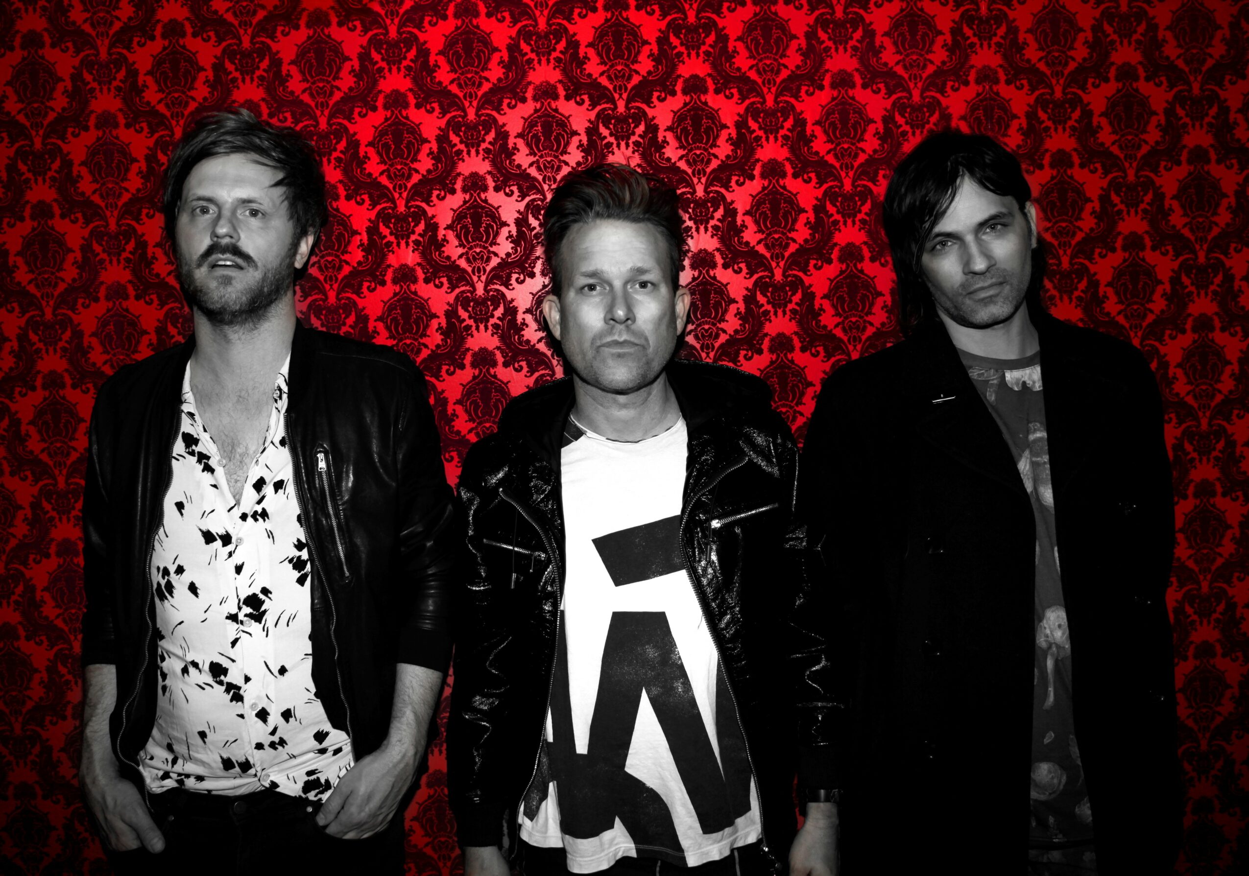 Three men stand in front of a red patterned background, wearing dark clothing and looking at the camera.