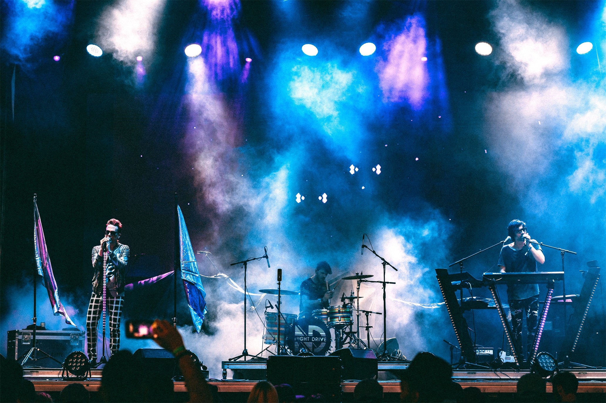 Stage with musicians performing, colorful lights, and smoke effects, with equipment and flags visible in the background.