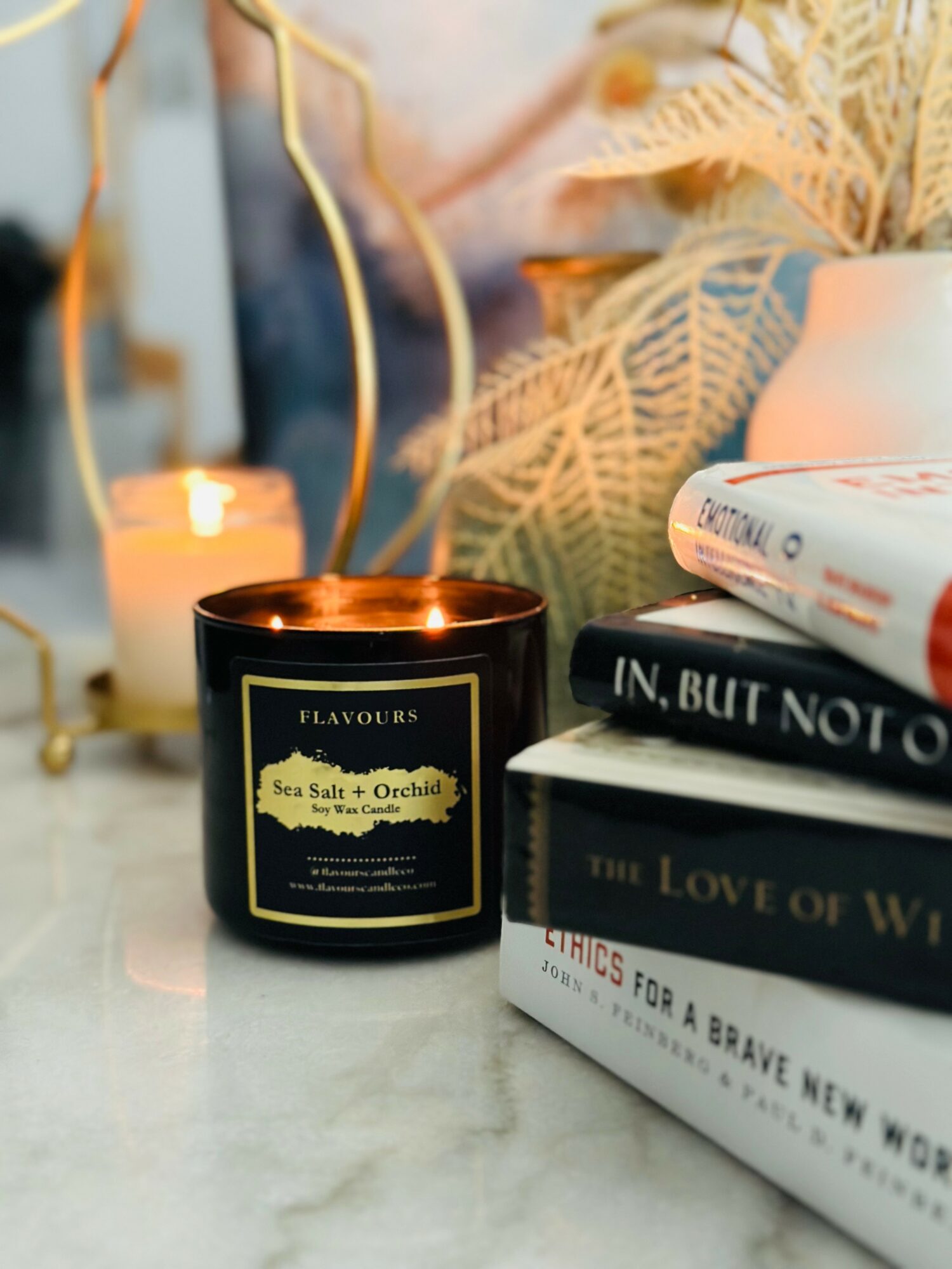 A lit black candle with a gold label, surrounded by books, a white vase, and decorative plants on a marble surface.