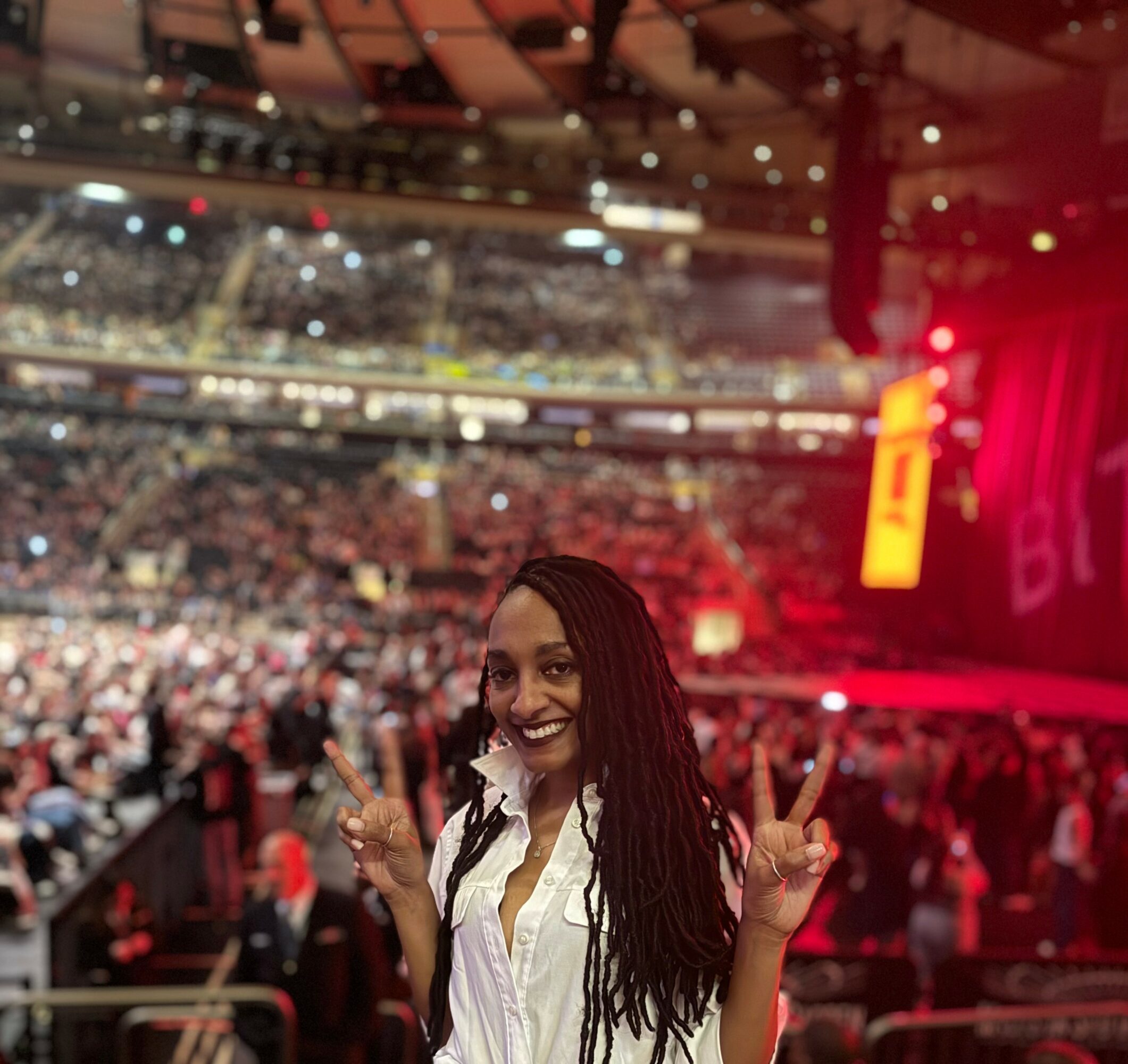 Smiling woman with long braids making peace signs at a crowded indoor event.