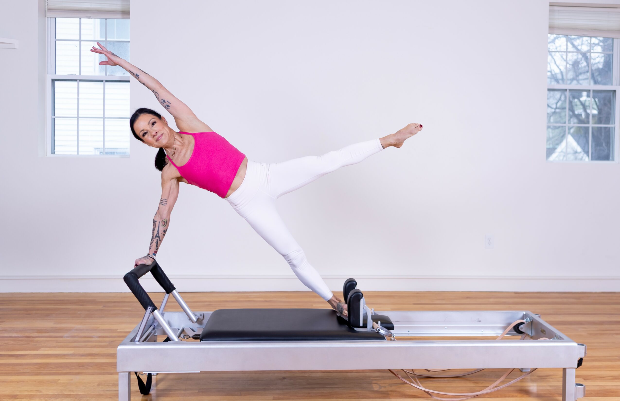 Woman in pink top and white pants performing a side plank on a Pilates reformer machine in a bright room with windows.