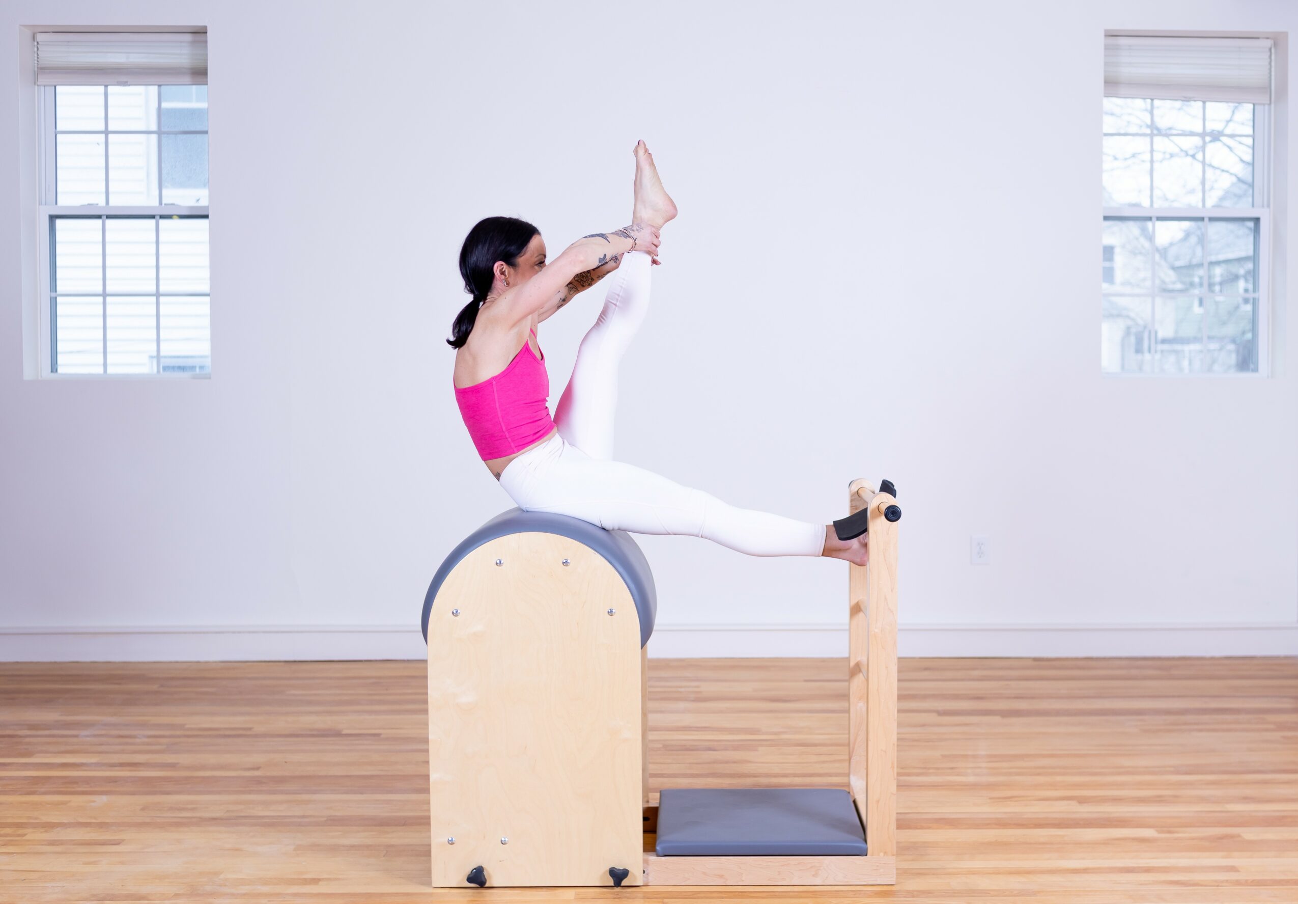 Person in pink top and white pants performing a stretch on a Pilates chair in a bright room with wooden floor and windows