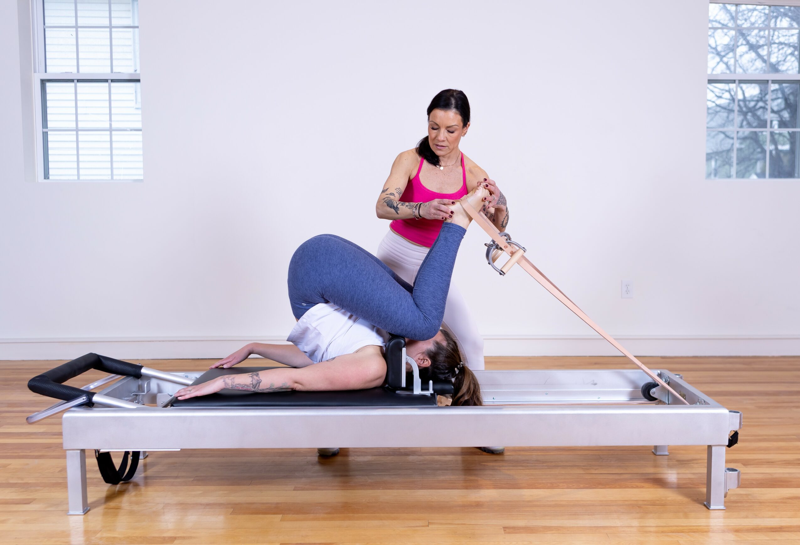 Person lying face down on a table, with another person standing beside, holding a strap attached to the person's leg, in a room with wooden floor and windows.