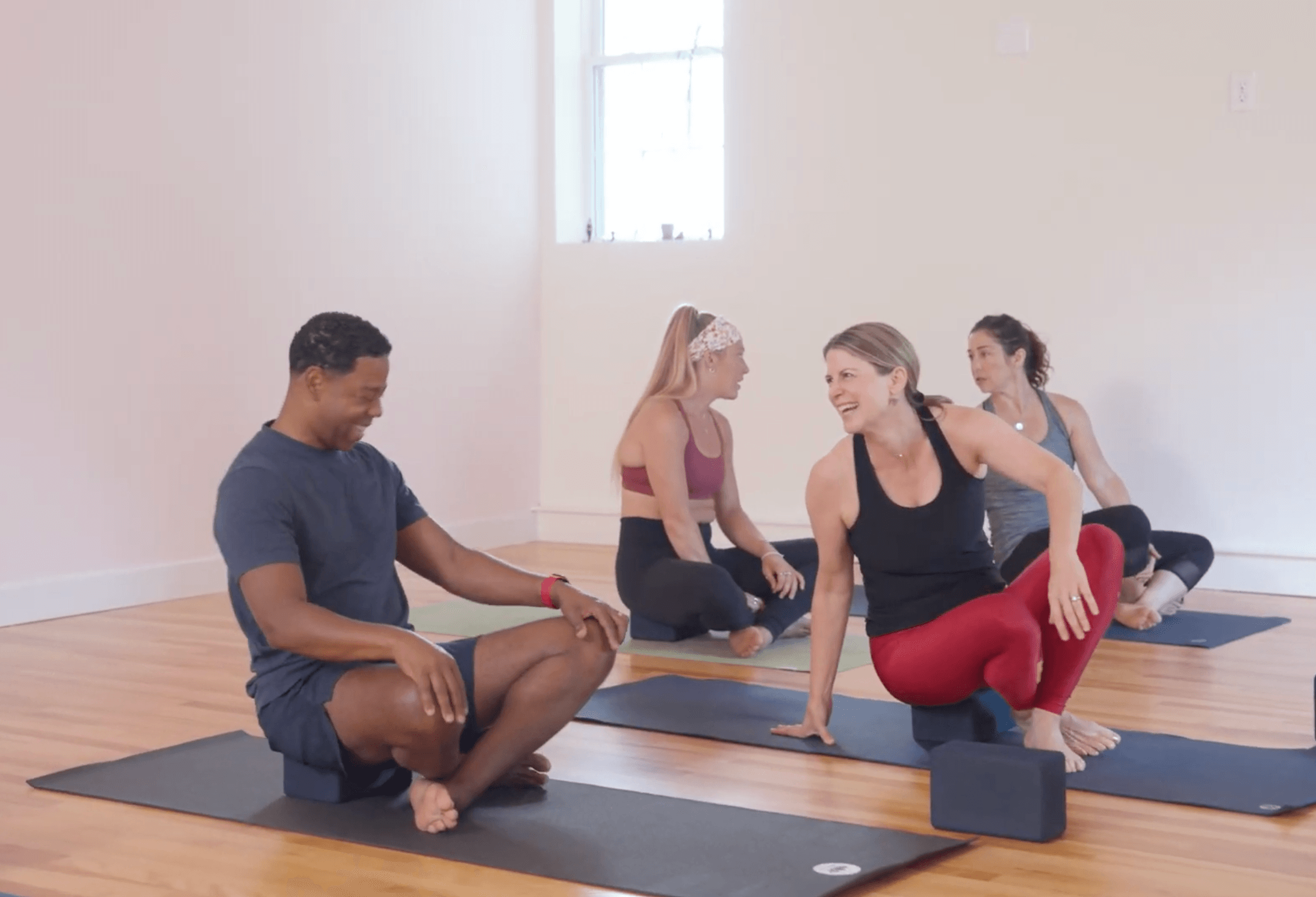 Four people in a yoga class sitting on mats in a bright room, engaging in conversation and stretching.