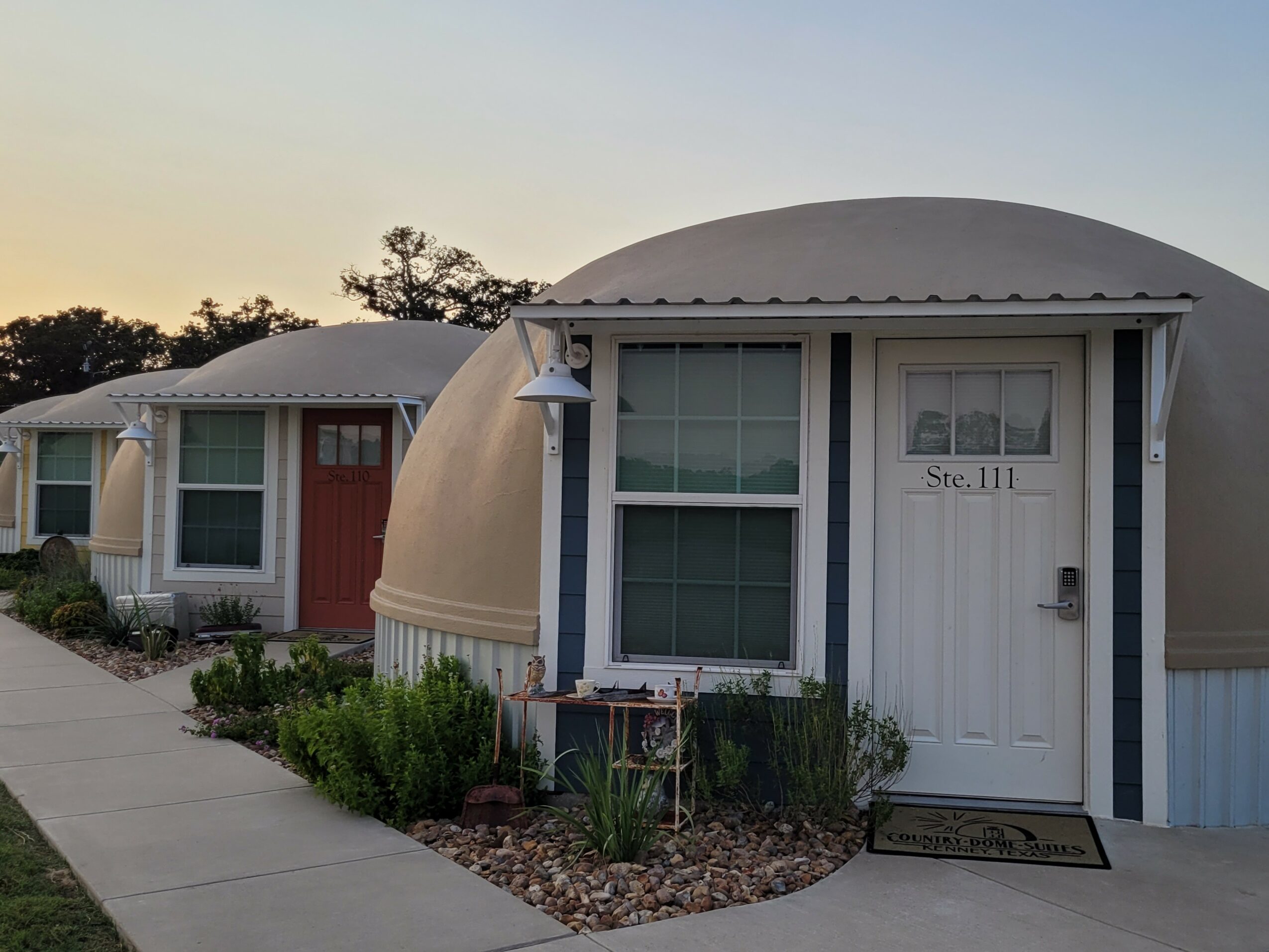 Small rounded house with white door and window, plants outside, and a lamp post, during sunset or sunrise.