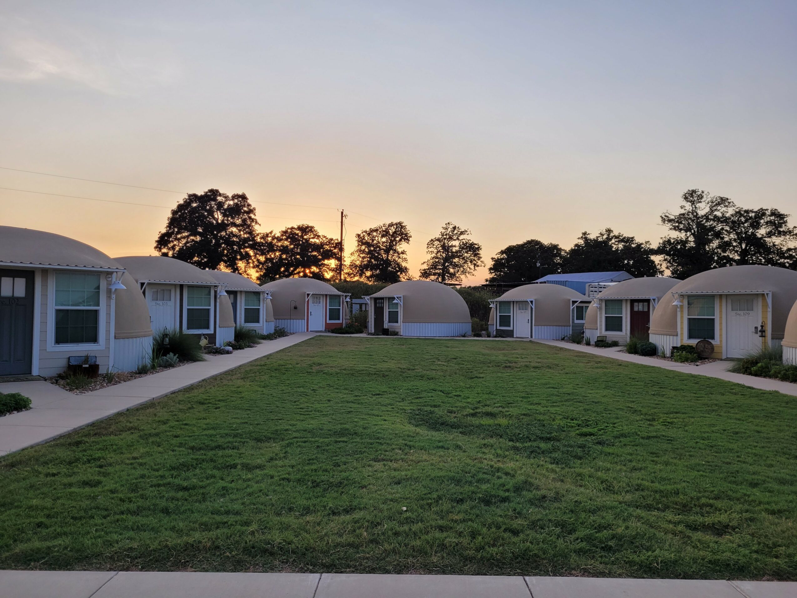 Row of small houses with front porches and a grassy area in front, trees in the background, sunset sky.