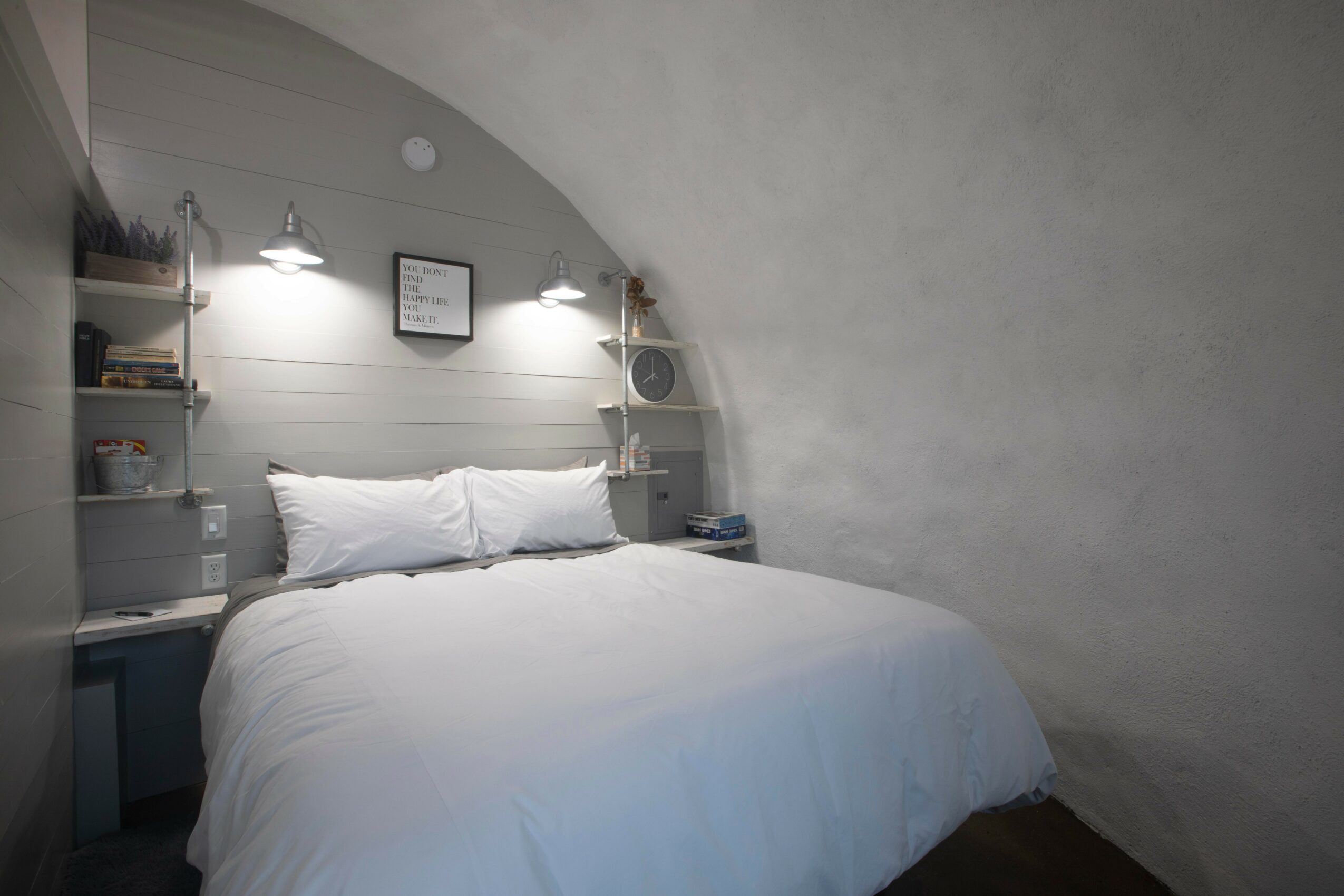 Bedroom with white bed, pillows, wall-mounted shelves, and wall lights, in a room with curved ceiling and gray wall.