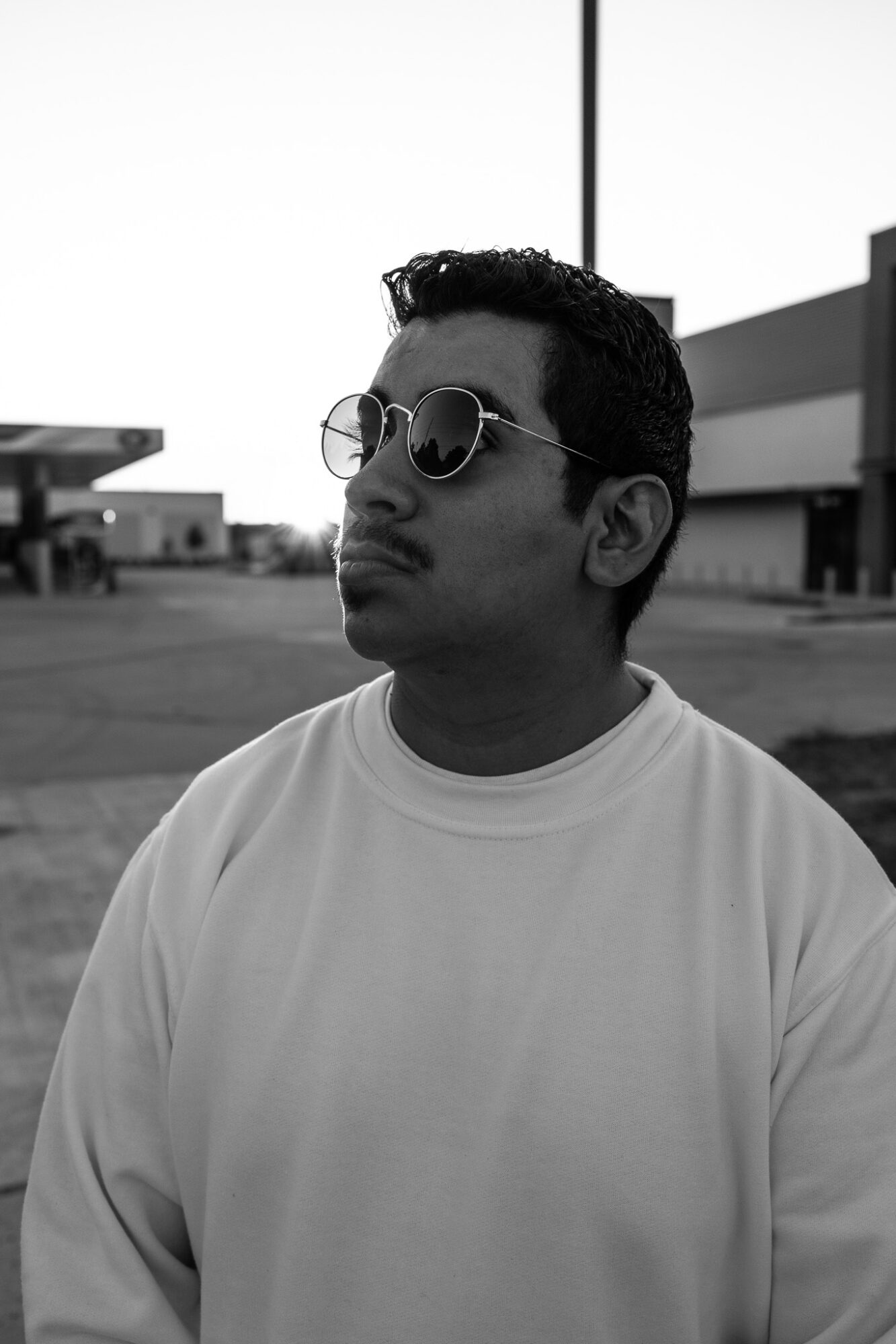 Young man wearing sunglasses and a light-colored shirt outdoors, with buildings and a pole in the background.