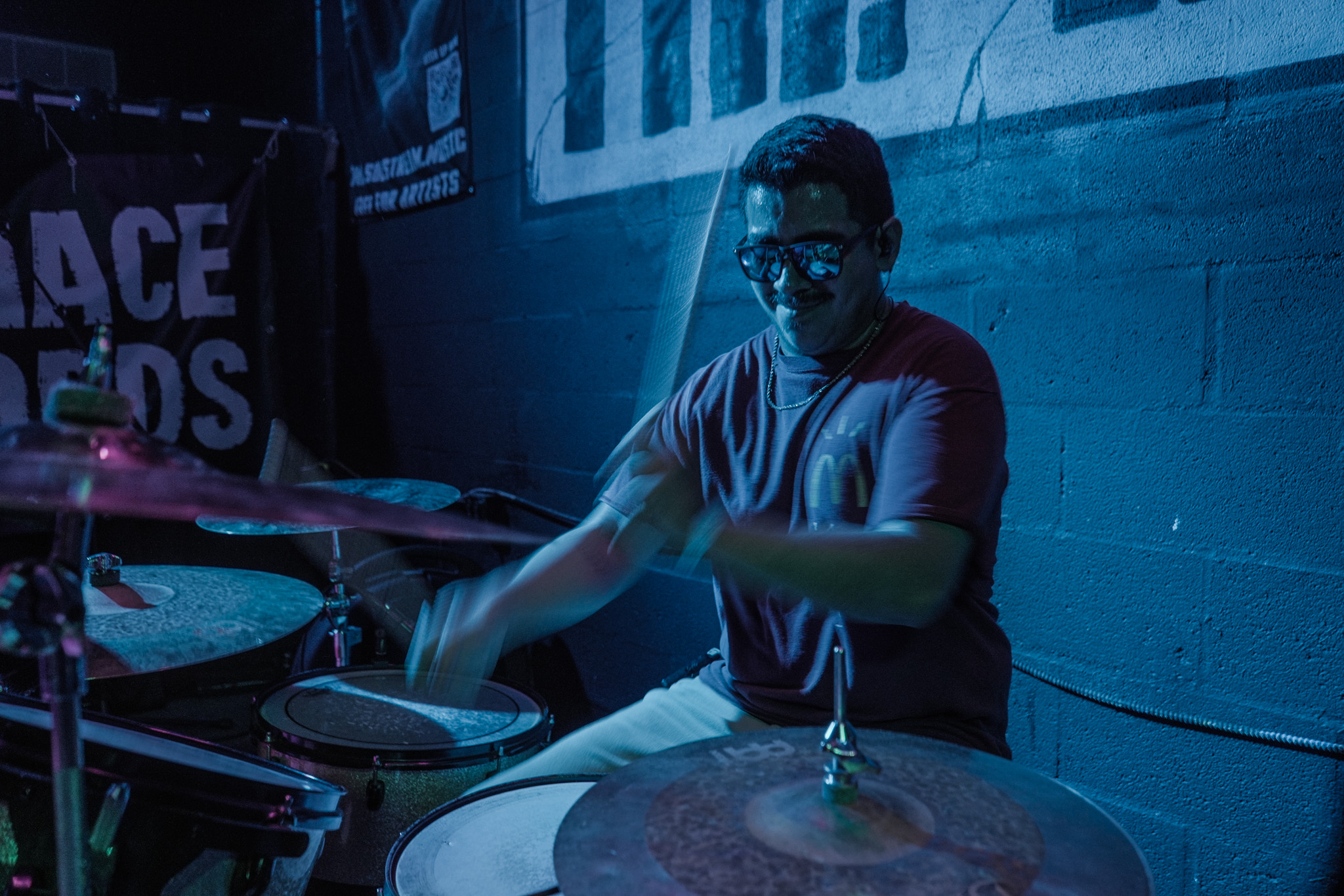 Person playing drums in a dimly lit room with blue lighting, wearing glasses and a t-shirt.