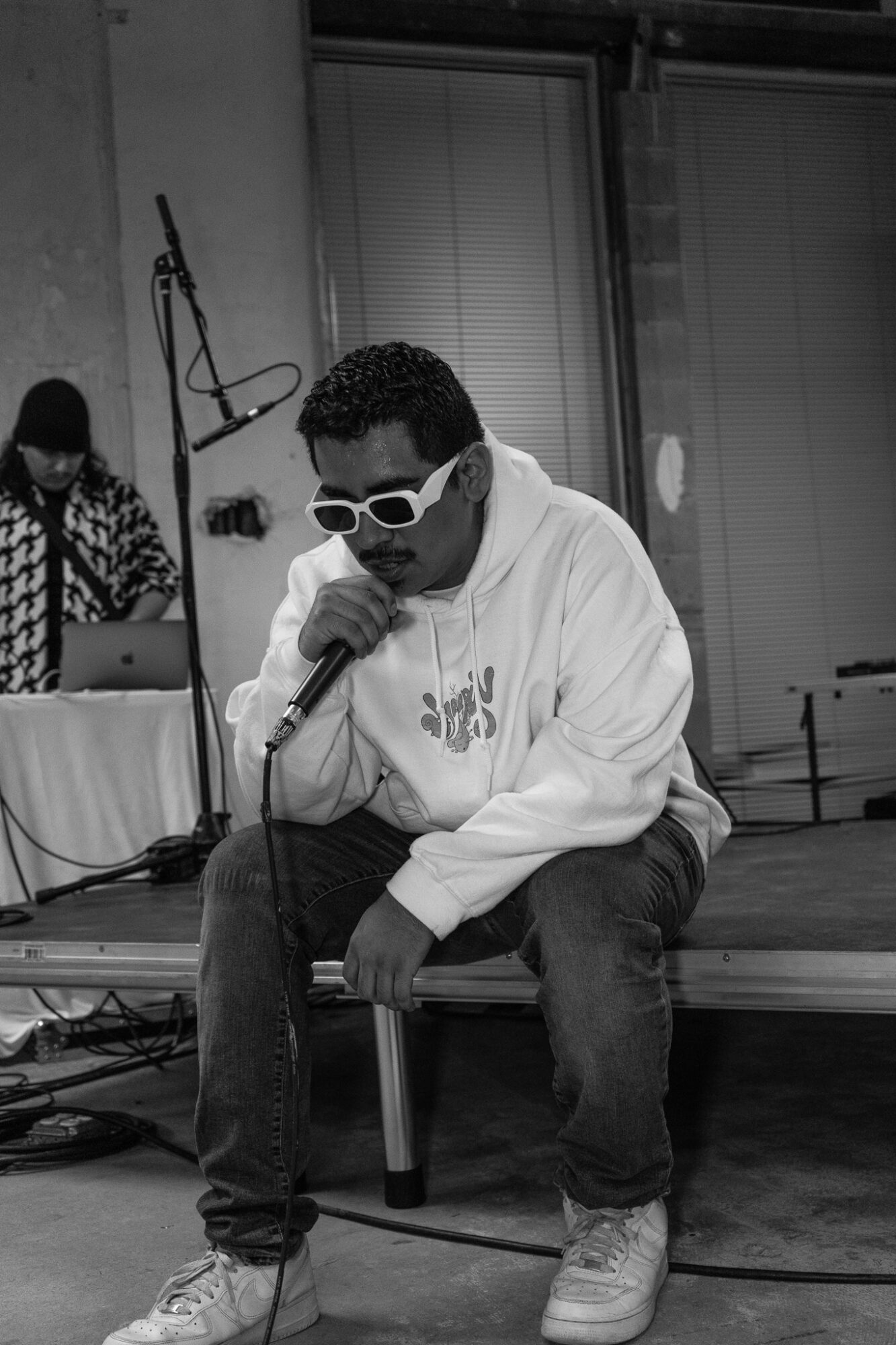 Person wearing sunglasses and hoodie, sitting on a stool, holding a microphone, performing indoors.