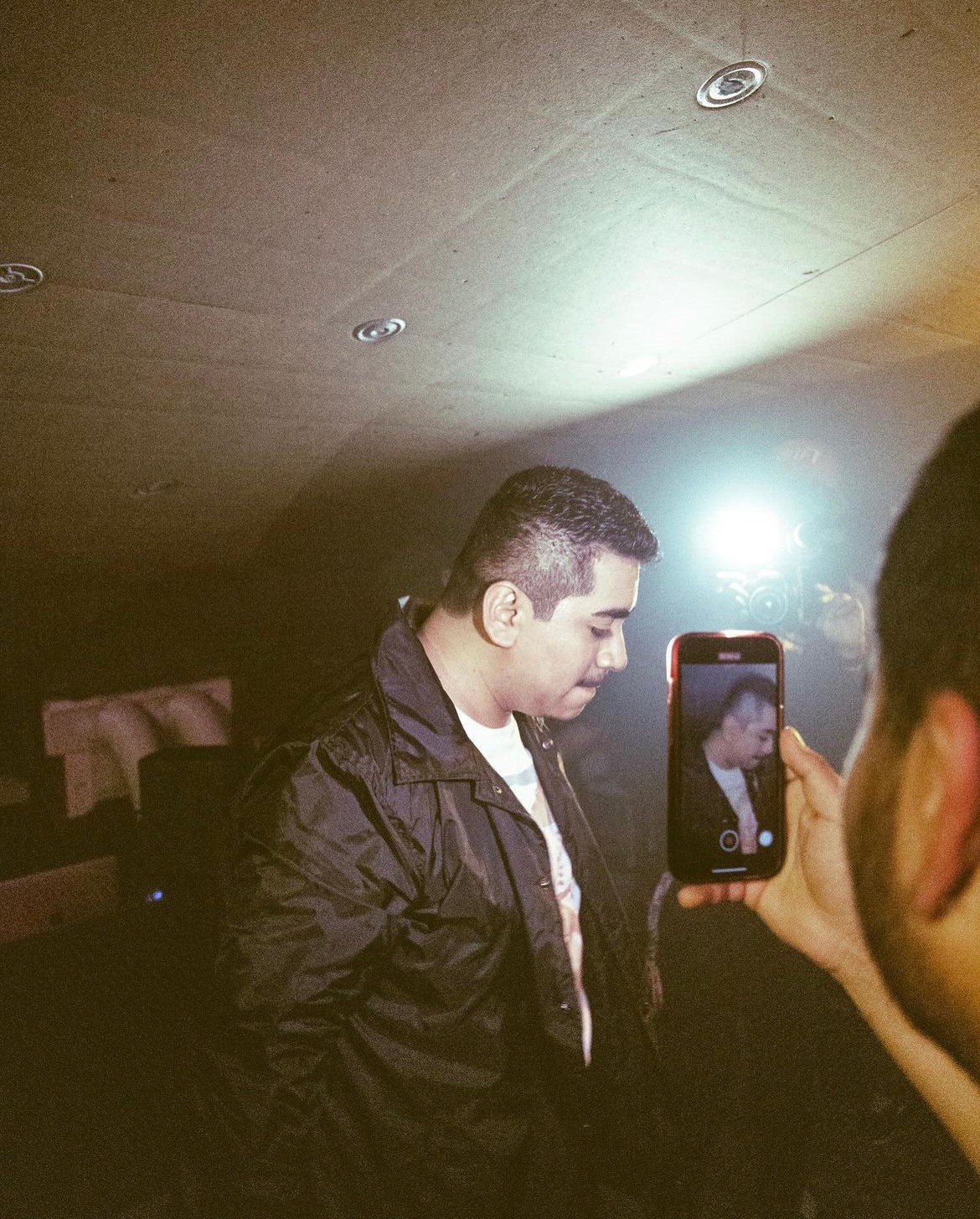 Person with short hair taking a photo of another person with a smartphone in a dimly lit room.